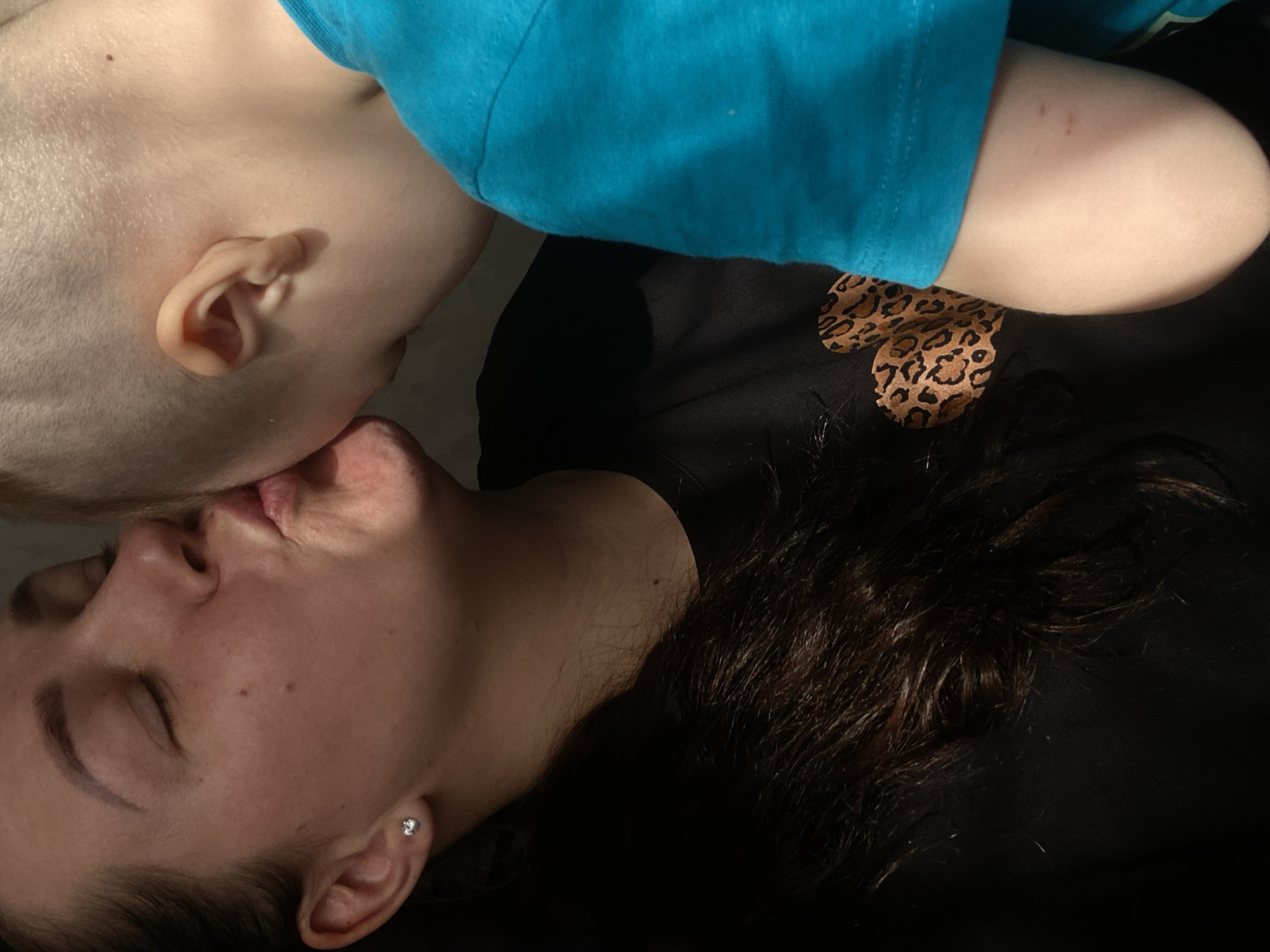 A close-up, high-angle shot captures a woman lying down while a child is kissing her. The woman is fair-skinned with dark hair and is wearing an earring. She appears to be wearing a black shirt with a leopard print design. The child, who is also fair-skinned, is wearing a blue shirt. The background is dark. The location is not discernible from the image.