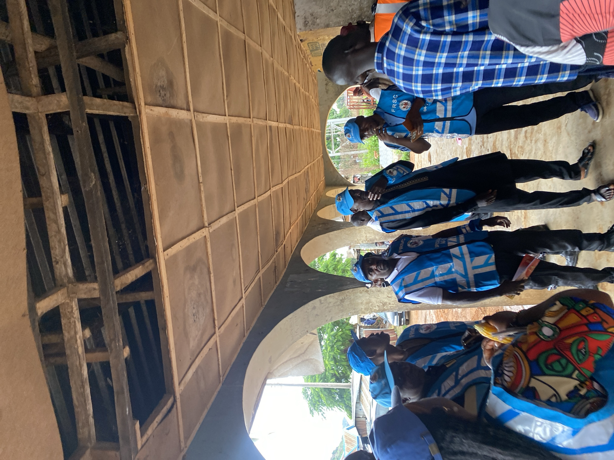 In Riyom, Nigeria, a group of individuals, many wearing matching blue caps and vests with reflective stripes, are gathered under a covered walkway. The ceiling of the walkway appears to be constructed of wooden beams and square panels, with some sections open to reveal the exposed roof structure. The individuals are of various ages and ethnicities, and some are engaged in conversation. One person is holding a piece of corn. The environment suggests an outdoor or semi-outdoor setting, possibly a market or a public gathering space, with glimpses of trees and buildings in the background. The overall atmosphere seems to be one of community engagement or a planned event.