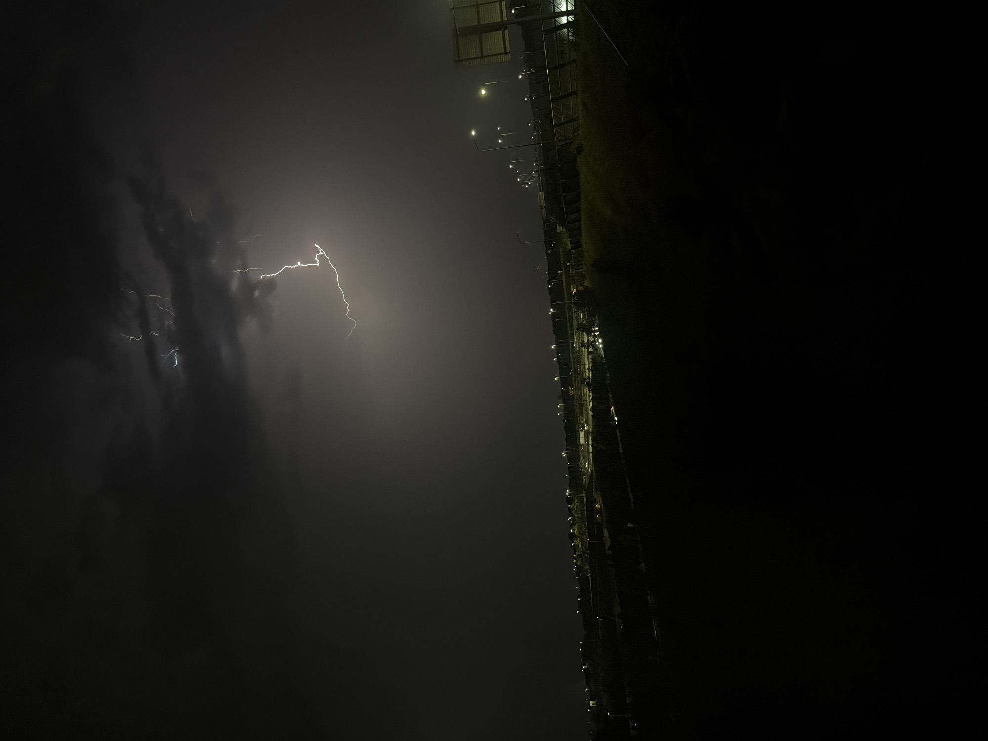 A nighttime scene captures a dramatic lightning strike illuminating dark, ominous storm clouds above a sprawling suburban landscape. The lightning forks across the sky, casting a bright, sharp light on the clouds and a portion of the horizon. Below, a dimly lit cityscape stretches out, characterized by rows of streetlights casting a warm glow on the empty roads and buildings. The foreground is dominated by dark, silhouetted hills and vegetation, suggesting a slightly elevated vantage point overlooking the illuminated area. The overall impression is one of a powerful natural event occurring over a quiet, unsuspecting urban environment. This appears to be a view of Badhoevedorp, Netherlands, given its characteristic flat terrain and the presence of numerous streetlights along what could be major roadways or developed areas. There are no people or discernible human activity visible in the image.