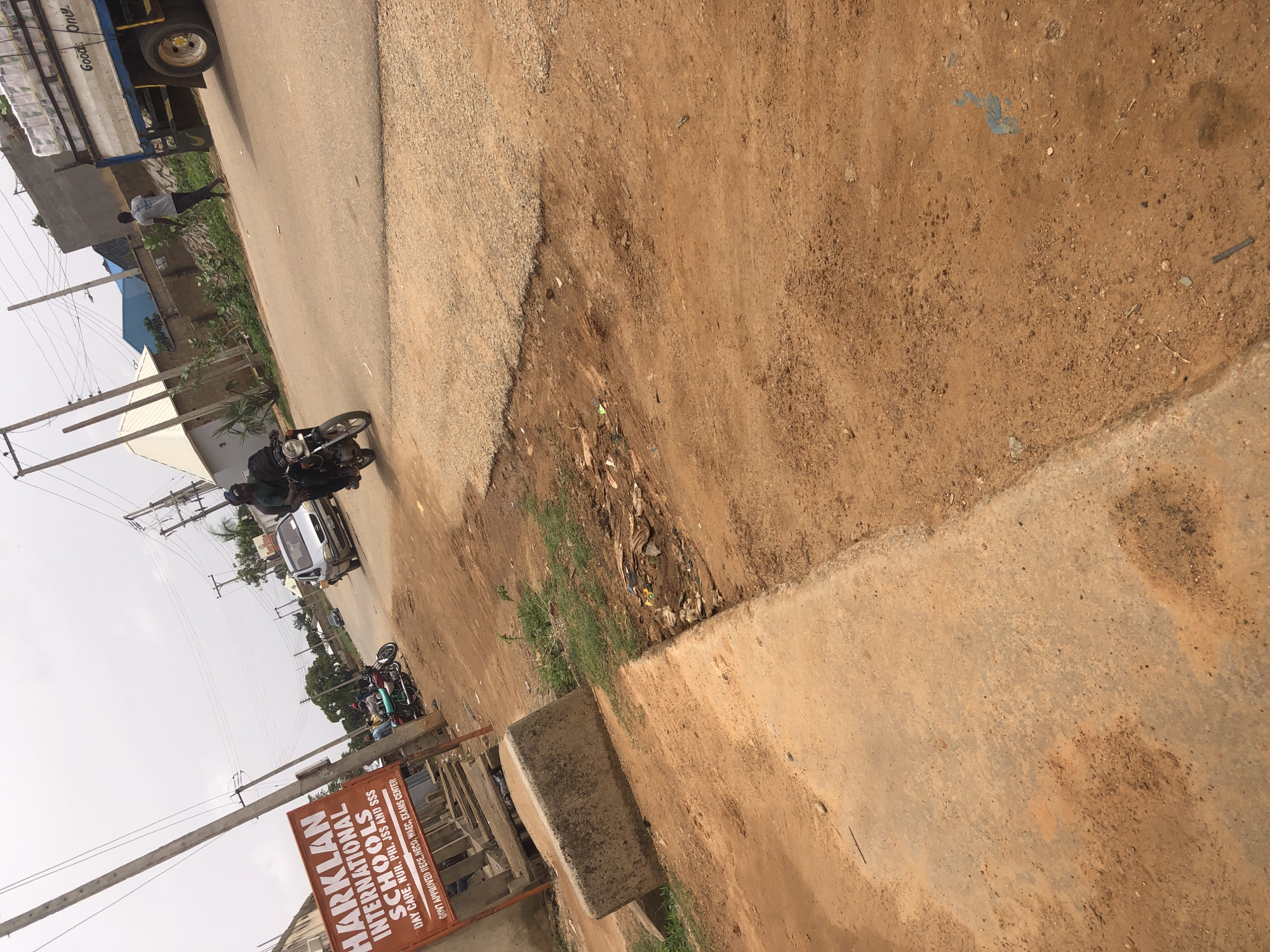 The image shows a street scene in Hanwa, Nigeria. A large truck is parked on the left side of the road, next to a man walking along the roadside. A motorcycle is also on the street. A sign for Harklan International Schools is visible near the bottom of the frame. The road is a mix of paved and unpaved surfaces, with dirt and debris along the edges. Overhead power lines are visible.