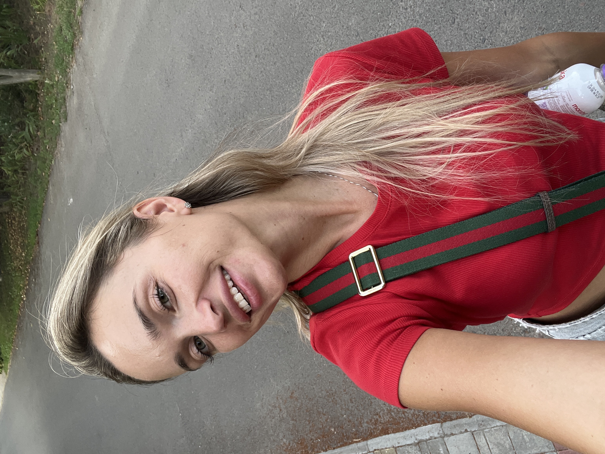 A young blonde woman with fair skin and green eyes is taking a selfie outdoors. She is wearing a red ribbed short-sleeved shirt and light-colored denim shorts. A green and red striped strap with a gold buckle is visible across her chest, likely from a bag. Her blonde hair is long and wavy, falling over her shoulders. She is smiling, showing her teeth. She is standing on a paved surface next to a sidewalk with some greenery and trees in the background. The context of Fontanka, Ukraine is not evident from the image itself. A bottle of liquid is visible on the right side of the image, partially obscured by her hair.