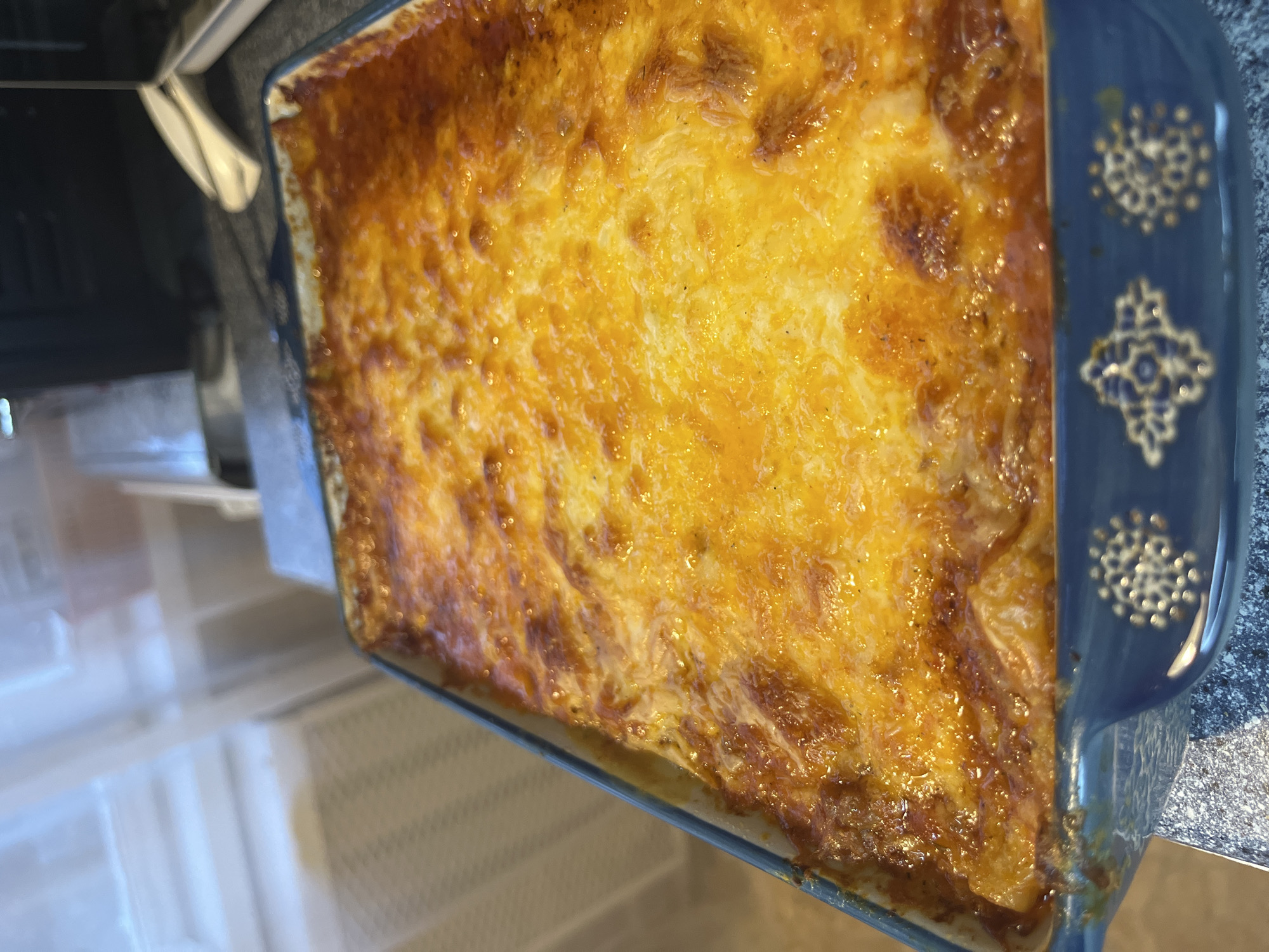 A baked dish, likely macaroni and cheese or a similar casserole, sits in a blue baking dish with decorative designs on the edges. The dish is golden brown and bubbly, suggesting it has been freshly baked. The location cannot be determined from the image.