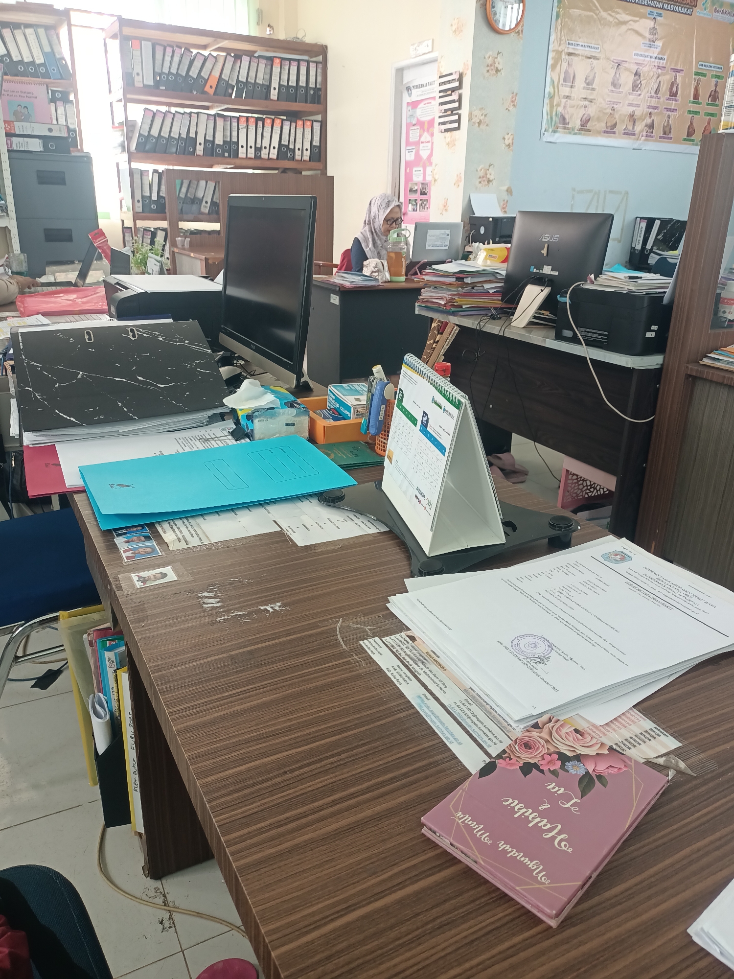 The image depicts an office setting in the city of Limbung, Indonesia. The scene is a close-up view of a wooden desk cluttered with various office supplies and documents. On the desk, there are several stacks of papers, files, a desk calendar, a computer monitor, and a printer. A light blue file folder is prominently placed on top of a stack of papers. To the right of the desk, a person wearing a headscarf and glasses is partially visible, appearing to be working at their computer. The background reveals bookshelves filled with binders and other office equipment, suggesting a busy administrative environment. The overall impression is of a functional, albeit somewhat disorganized, workspace.