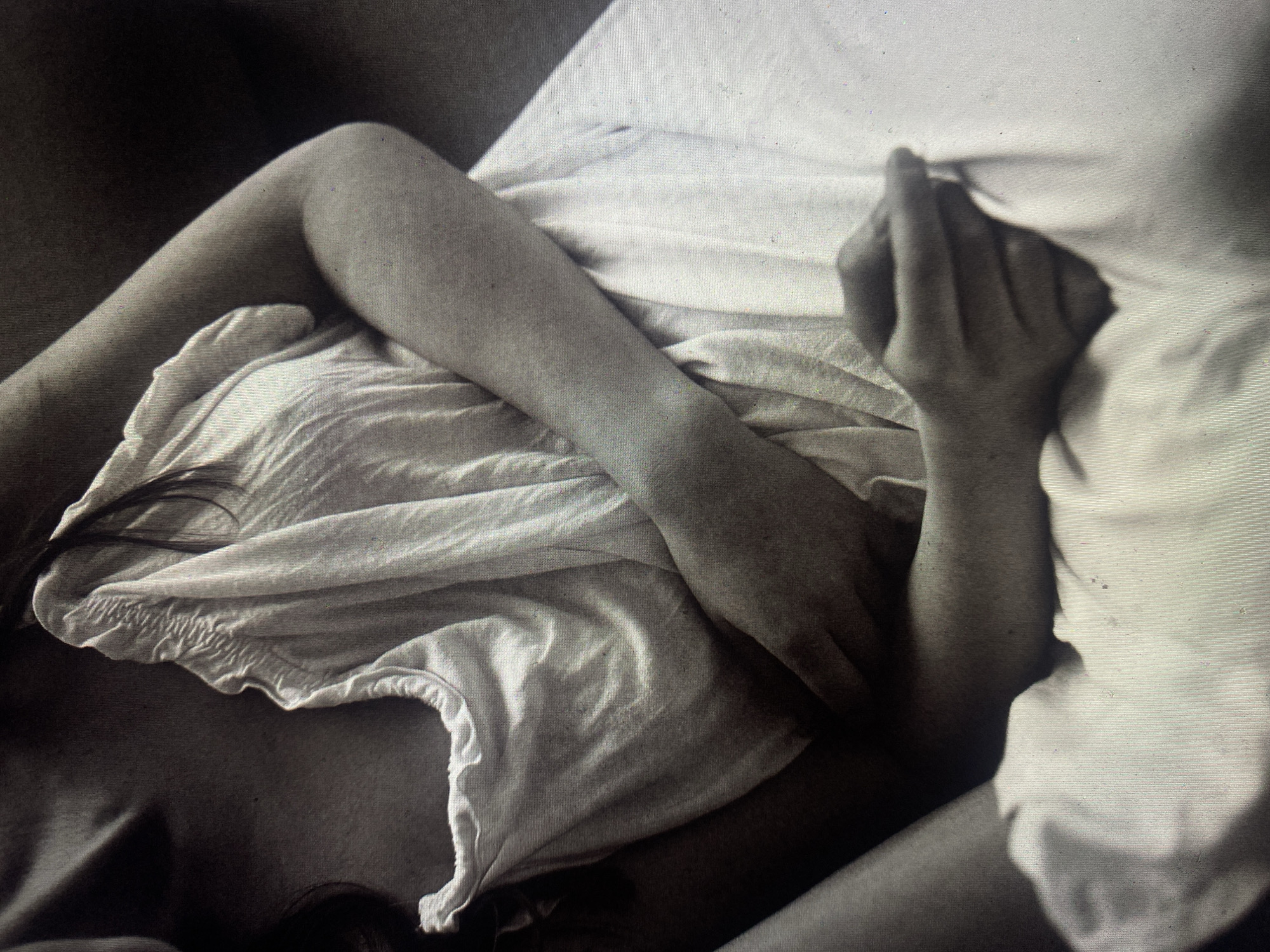 A monochrome photograph shows a person lying in bed. A leg is visible in the upper left of the frame, with a white, loose-fitting garment (possibly nightwear) draped over the torso. The right arm extends across the bed, the hand gently gripping or touching the white bedsheet. The person's head and hair are partially visible in the lower center of the frame. The setting appears to be indoors, and there is no evidence of location in the city of Шебунино, Russia.