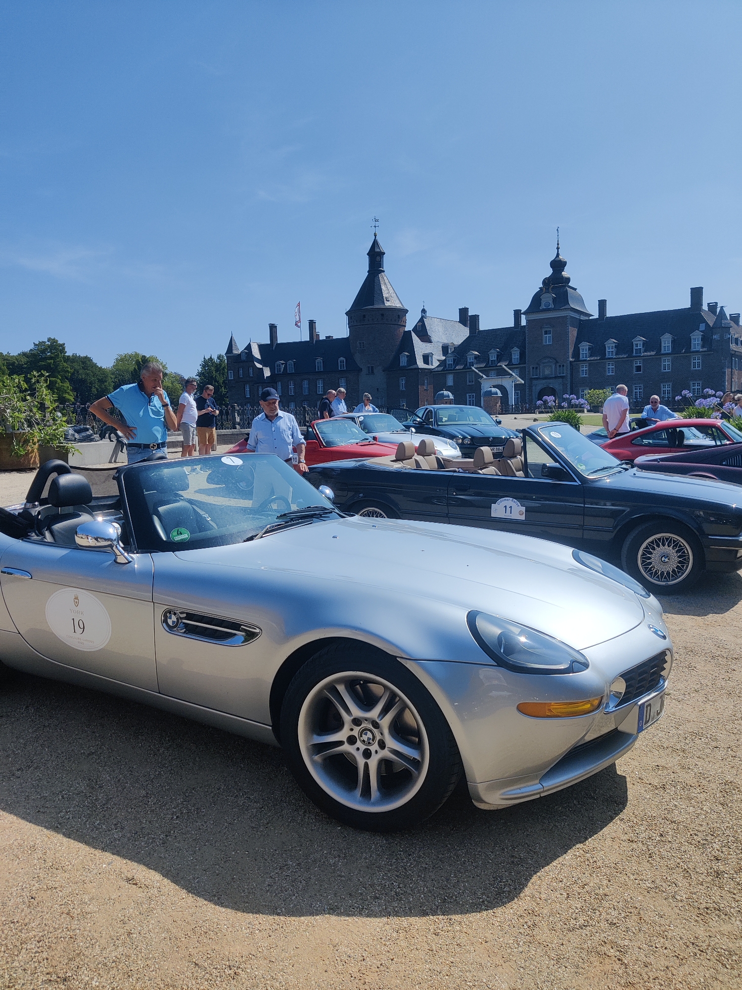 The image captures an outdoor event with several classic cars parked on a gravel surface in front of a large, ornate castle under a clear blue sky. In the foreground, a silver convertible BMW Z8 is prominently displayed, showing its sleek design and a white oval sticker with the number "19" on its side. Behind it, a dark-colored convertible is visible, with a sticker displaying the number "11". Further back, a red convertible and a dark sedan are parked. Several people are standing around the cars, some appearing to admire them. The castle in the background, likely Wasserschloss Anholt, is a prominent feature of the scene, with its distinctive architecture and multiple towers. The overall atmosphere suggests a car show or a similar gathering of automotive enthusiasts at a historic location.