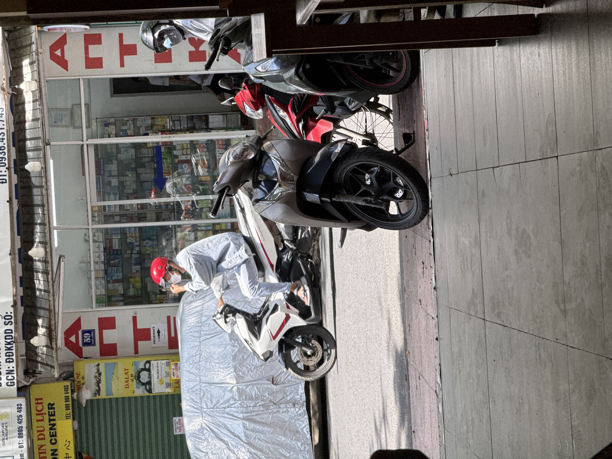 A street scene in Nha Trang, Vietnam, features a pharmacy with a prominent red "A" logo. In front of the pharmacy, a person wearing a red helmet and a light-colored jacket rides a white and red motorcycle. Another individual in the background wears a helmet with a face shield. Several other motorcycles are parked on the sidewalk. The storefront displays shelves stocked with various products, and a sign with the number "59" is visible. Sunlight casts shadows on the street and sidewalk.