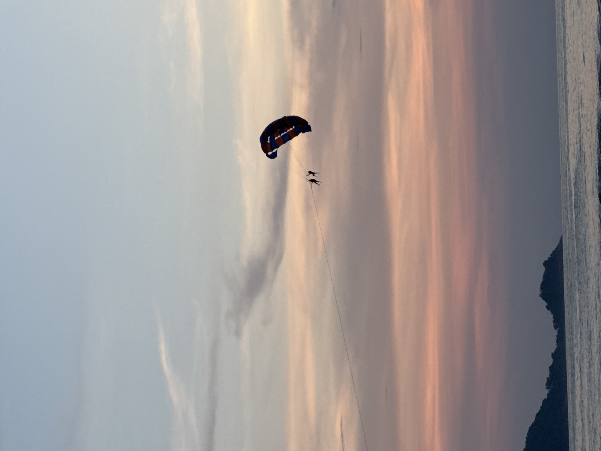 The image captures a parasailing activity at sunset over the ocean in Patong, Thailand. Two individuals are suspended in the air, attached to a colorful parasail with dark blue and red accents, being pulled by an unseen boat. The sky displays a gradient of soft hues, transitioning from pale blue at the top to shades of pink and orange, indicative of a sunset. Wispy clouds are scattered across the sky. In the foreground, the calm ocean surface reflects the sky's colors, and the dark silhouette of a hilly coastline, likely part of Patong, is visible on the left side of the frame.