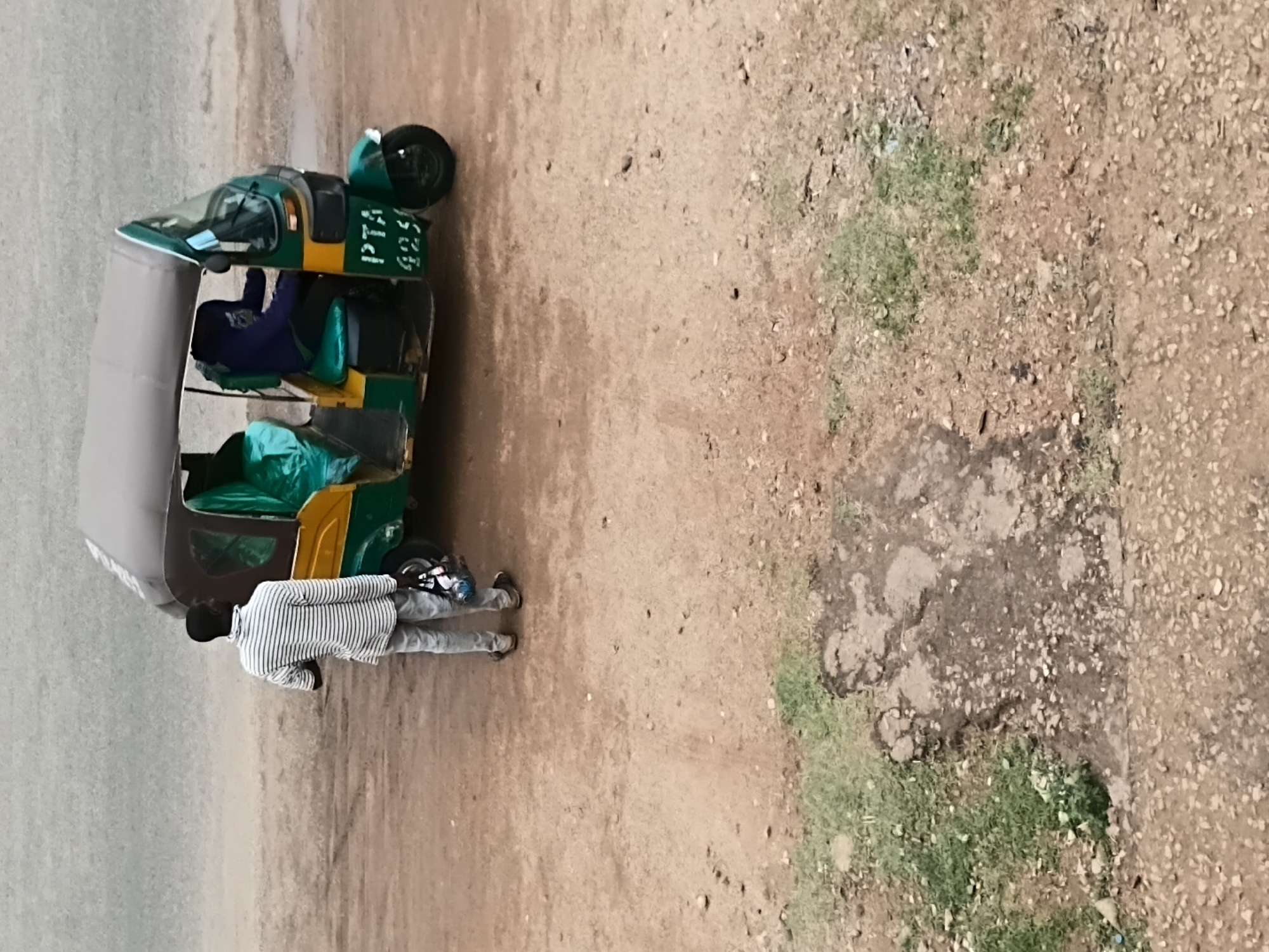 The image shows a three-wheeled motorized vehicle, commonly known as a "keke" or auto-rickshaw, parked on a dirt road in what appears to be a semi-urban area. A man wearing a striped shirt is standing near the back of the vehicle. Another individual is sitting inside the keke. The vehicle is green, yellow, and grey. The ground is mostly dirt and gravel with patches of sparse green vegetation. Based on the style of the vehicle and general environment, it is likely in Jos, Nigeria. The scene appears to be outside.