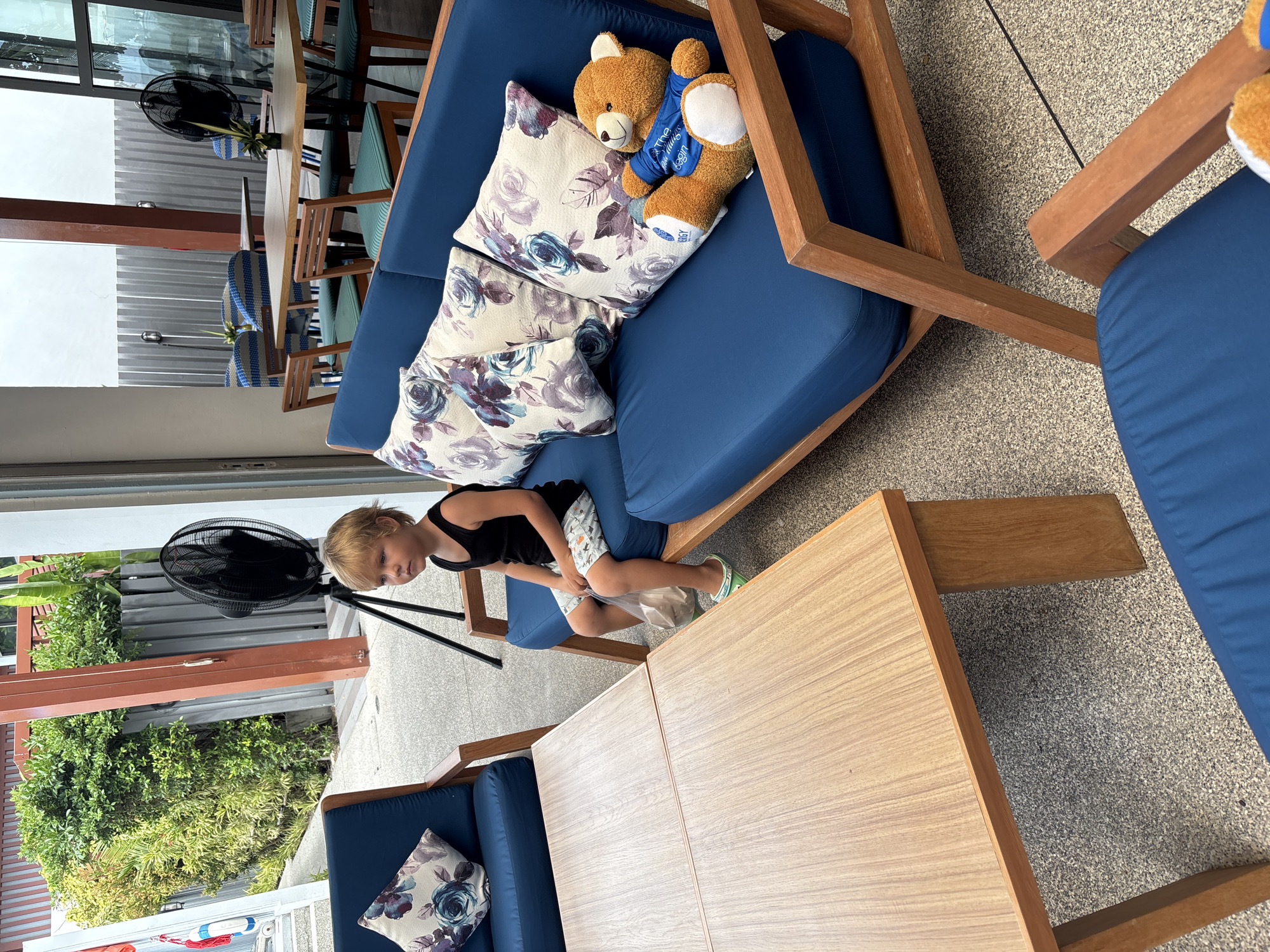 The image shows an outdoor seating area, likely at a restaurant or cafe, in Baan Chaweng, Thailand. A young boy with blond hair, wearing a black tank top and patterned shorts, sits on a blue outdoor sofa. He is holding a plastic bag in his left hand and wearing green and white sandals. A stuffed teddy bear in a blue shirt is also seated on the sofa next to him. In front of the sofa is a light-colored wooden coffee table. The background shows more outdoor seating with tables and chairs, and lush green foliage. A black oscillating fan stands behind the sofa. The flooring appears to be a textured grey material, possibly concrete or asphalt. The overall atmosphere suggests a relaxed, possibly tropical, setting.