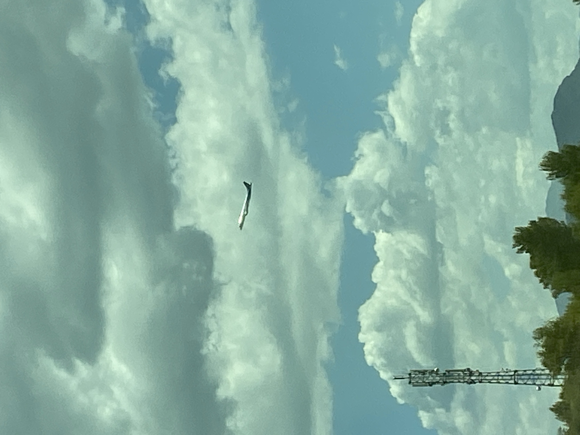 A distant, white airplane with a dark tail is captured in flight against a partly cloudy sky. The clouds are a mix of light gray and white, with patches of blue sky visible. In the foreground, a tall, metal telecommunications tower stands prominently on the left side of the frame. Below the tower and to the right, the tops of green trees are visible, suggesting a natural landscape. In the far background, the faint outline of a mountain range can be seen against the horizon. The image appears to be taken during the daytime under fair weather conditions.