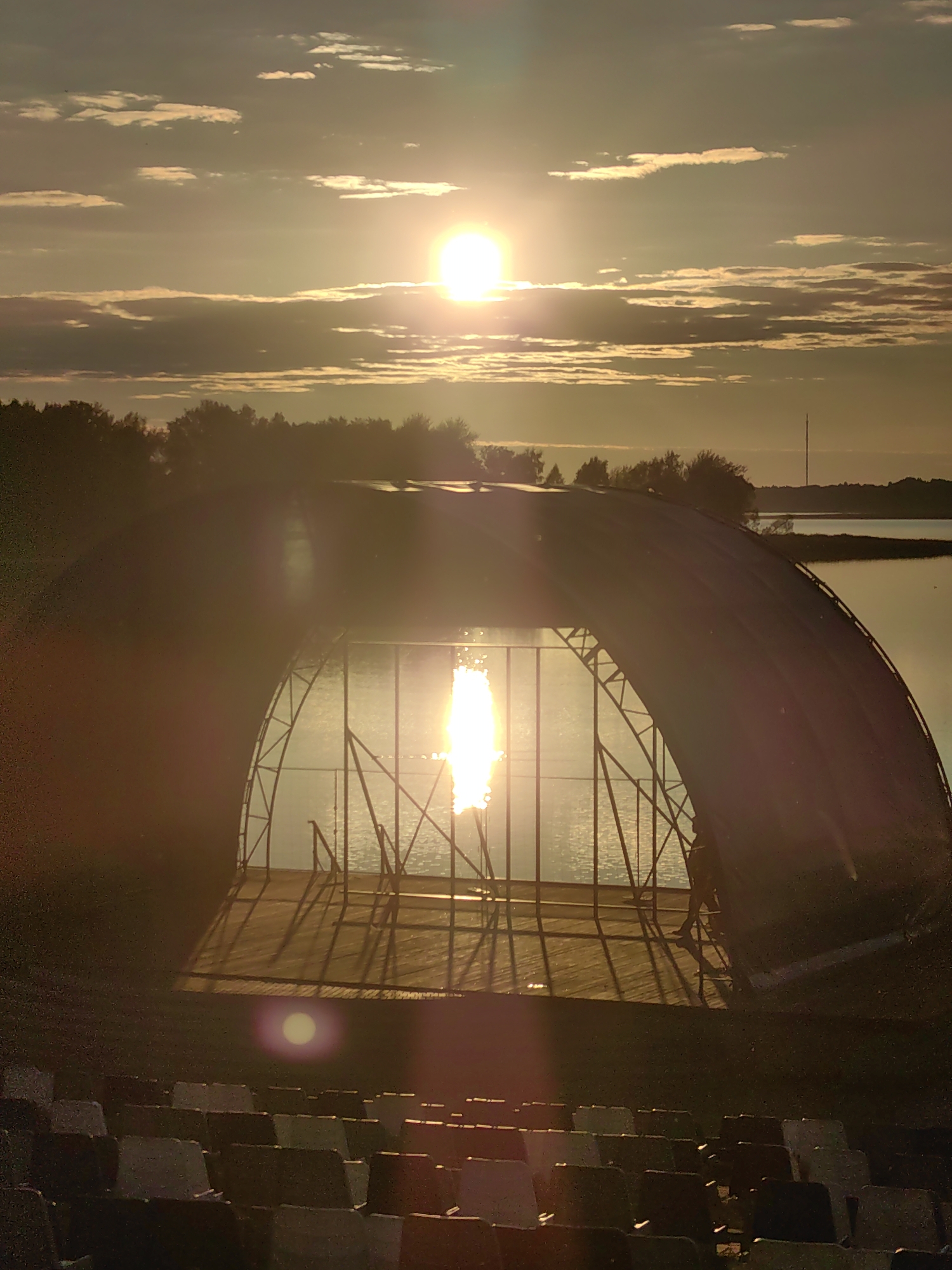An outdoor amphitheater is pictured at sunset in Myadzyel, Belarus. The sun, a bright orb with rays emanating from it, is positioned in the upper center of the frame, partially obscured by clouds. The light casts a warm glow over the scene. In the foreground, rows of empty seats are visible, some with the backs tilted up. Behind the seats, a large, open-sided structure with a curved roof stands. This structure appears to be a stage or performance area, with a wooden floor and metal support beams. The sun's reflection creates a bright, shimmering path across the water in the background, beyond the structure. The water is calm, with a tree line silhouetted against the horizon. A thin, tall tower is visible in the distance on the right side of the frame. The overall impression is one of tranquility and the anticipation of an evening event.