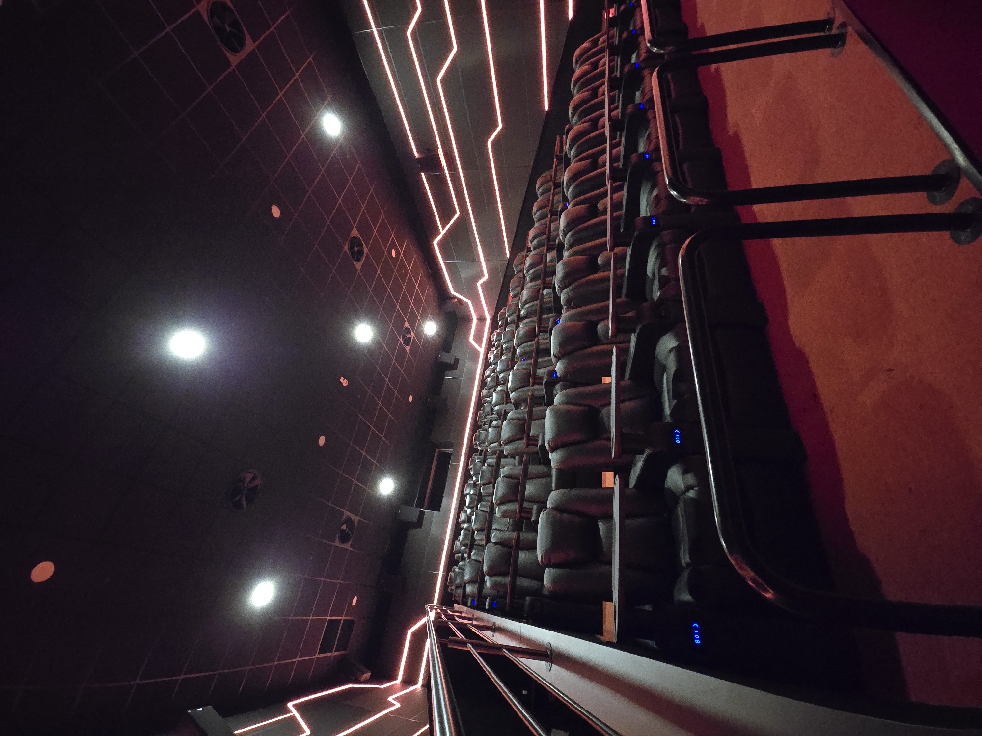 The image displays an unoccupied, dimly lit cinema auditorium, identified as a premium screen within Cinema City Varna at Delta Planet Mall, Varna, Bulgaria. The perspective is angled from the lower front-right of the room, looking upward and across multiple ascending rows of dark, individual, reclinable cinema seats. Each seat is equipped with a small, circular, swiveling side table. Blue LED indicators, such as "B01" and "B02", displaying seat numbers, are visible on the outer frame of some seats. A metal handrail system extends along an elevated walkway or aisle in the left foreground. The floor is covered in red carpeting. Dark walls are accented with horizontal, angular patterns formed by red LED strip lighting. The black ceiling features a grid pattern, incorporating multiple recessed circular white lights and circular ventilation grates. No individuals are present in the auditorium.