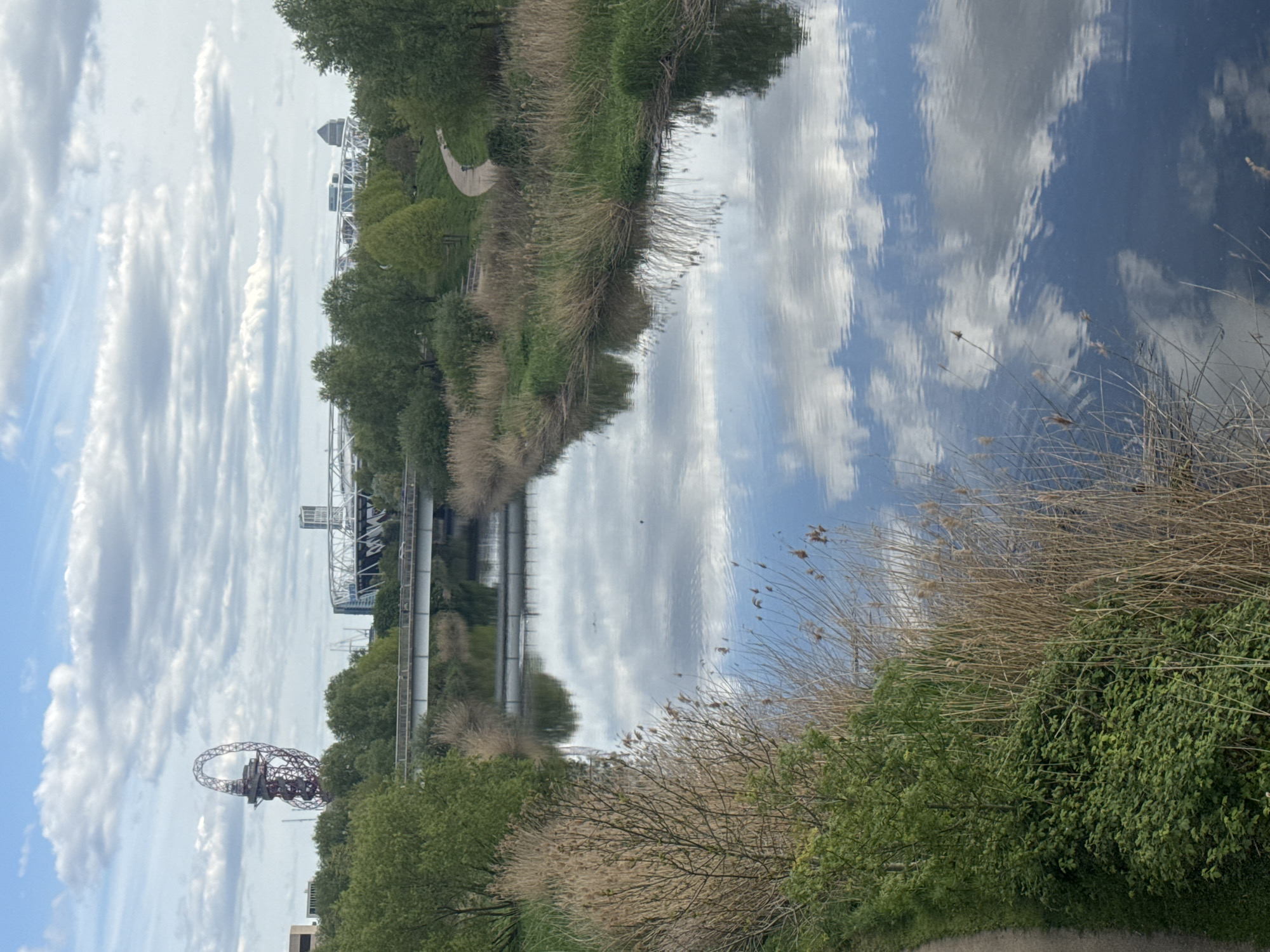 The image depicts a waterway running through Queen Elizabeth Olympic Park in London, United Kingdom. Along the banks are trees and reeds, reflecting in the water along with the sky. In the distance are views of the ArcelorMittal Orbit and the London Stadium. A path runs parallel to the waterway. The sky is partly cloudy.