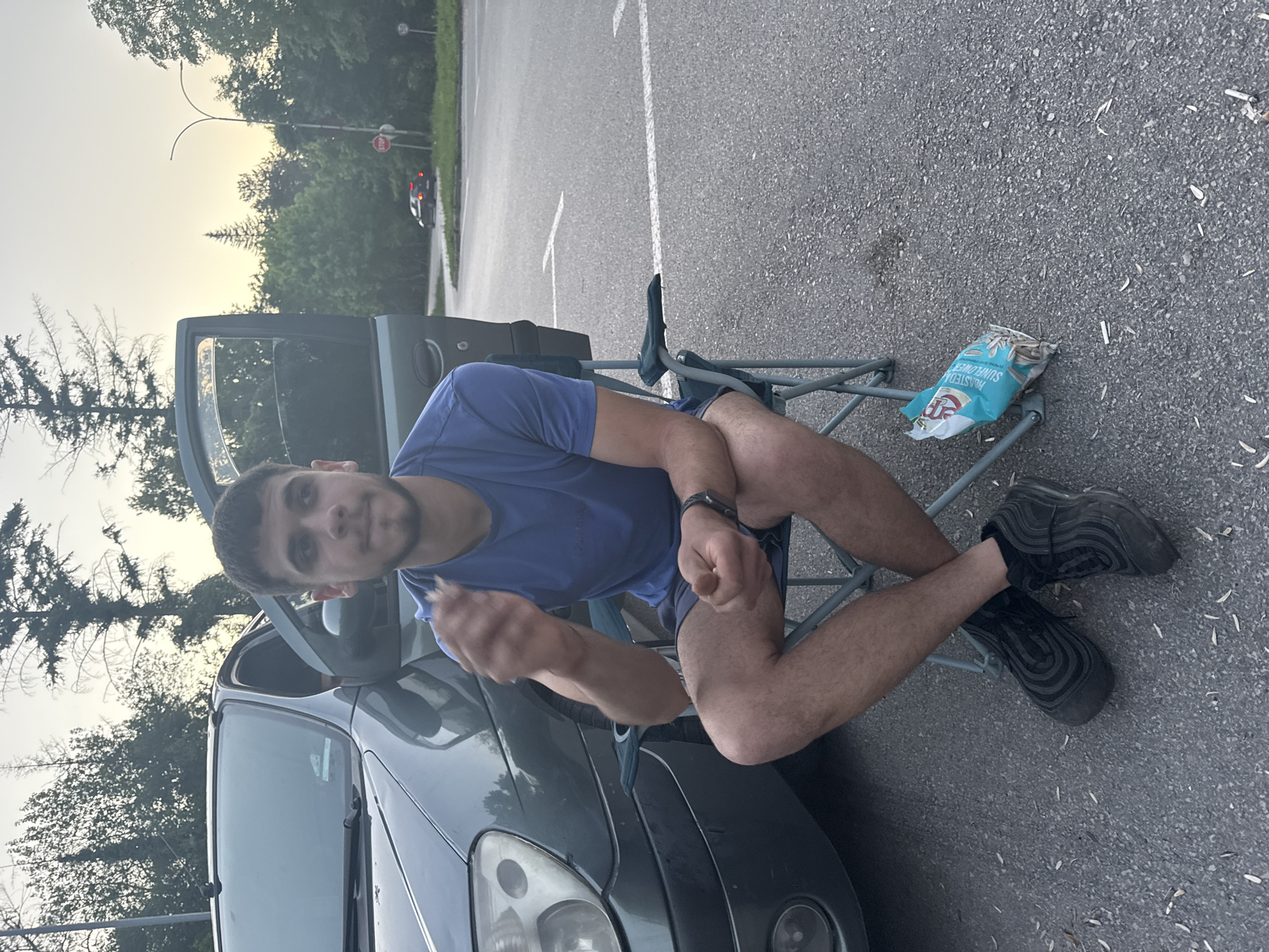 A man is sitting on a folding chair next to a parked car. The car door is open. There is a bag of sunflower seeds on the ground next to the chair. The scene is outdoors, in a parking lot with a road and trees in the background. A "Stop" sign is visible in the background. Location details for Pleven, Bulgaria are not discernible from the visual information provided.