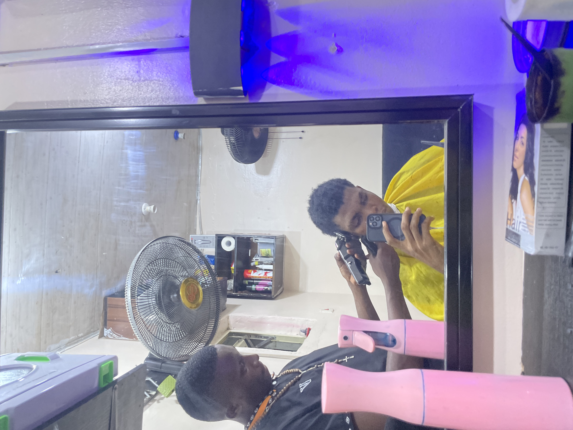 The image is a selfie taken in a mirror, reflecting the inside of what appears to be a barber shop, possibly in Ado, Nigeria. Reflected in the mirror is a man wearing a yellow barber cape, holding a phone in one hand and clippers in the other, seemingly taking a photo of himself getting a haircut. His head is partially shaven. A second man is visible in the mirror lying on a barber's chair, wearing a black shirt with a beaded necklace.
In the background are items commonly found in a barber shop, including a fan, a cabinet with products, and other supplies. On the right side of the mirror are a pair of sunglasses and a hair product advertisement. The environment suggests an establishment where grooming services are provided. The lighting in the space appears to be a combination of natural and artificial, with blue-toned artificial lights above the mirror. There are also two pink spray bottles reflected in the mirror, positioned in the lower foreground. The walls appear to be painted in neutral, pale shades.