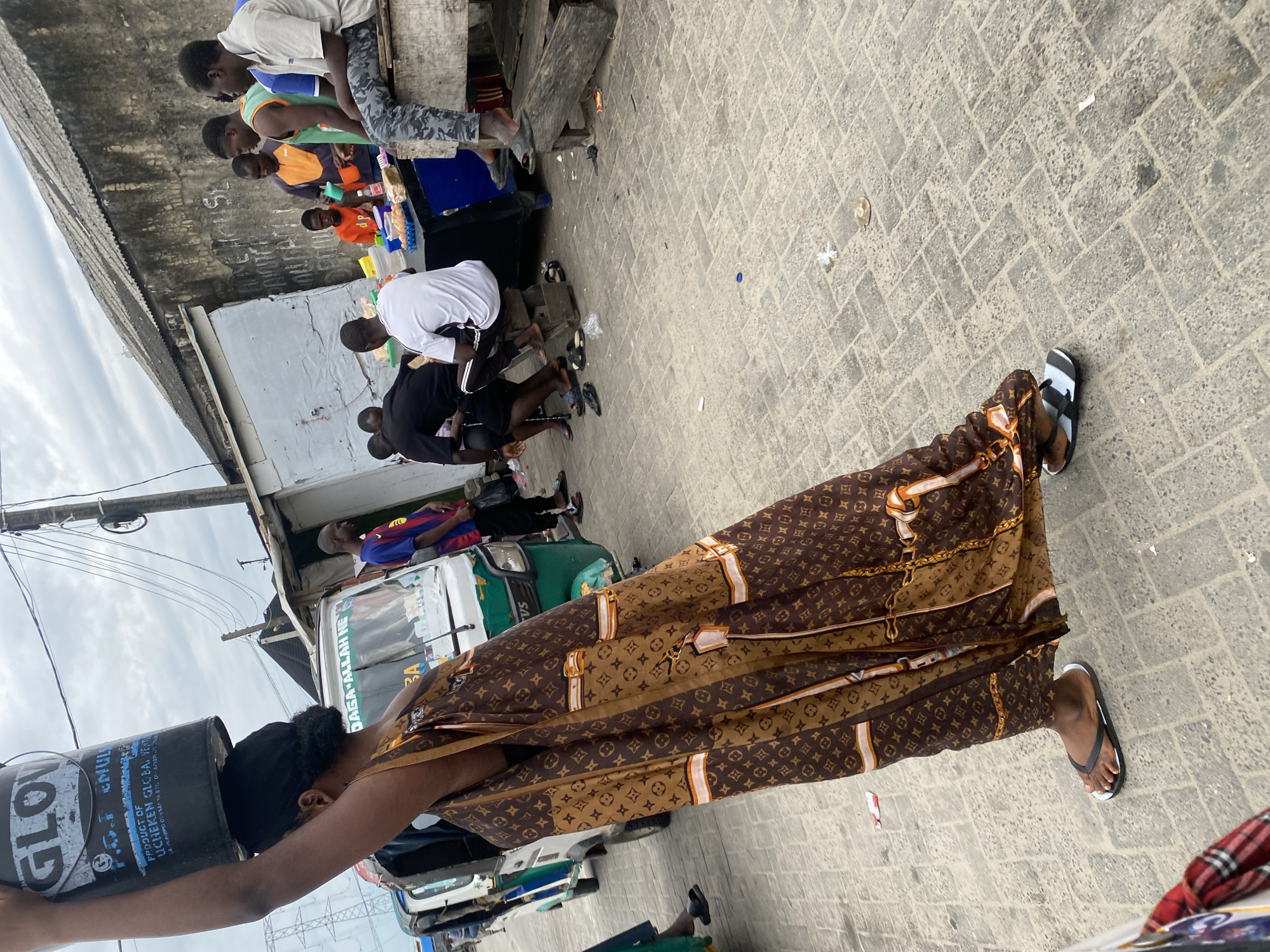 In Ado, Nigeria, a young Black woman balances a large black bucket on her head, its label indicating "GLOW." She is wearing a long, brown patterned dress with a Louis Vuitton-like design and black flip-flops. To her right, a white and green three-wheeled vehicle is parked. Further to the right, a group of young Black men are gathered around a makeshift stall. One man in an orange shirt is serving food from containers on a table. Other men are seated or standing nearby, some wearing athletic attire. The ground is paved with gray bricks. Overhead, a cloudy sky is visible with power lines and a utility pole. The background shows the weathered walls and corrugated roofs of several structures, suggesting an urban or semi-urban setting.