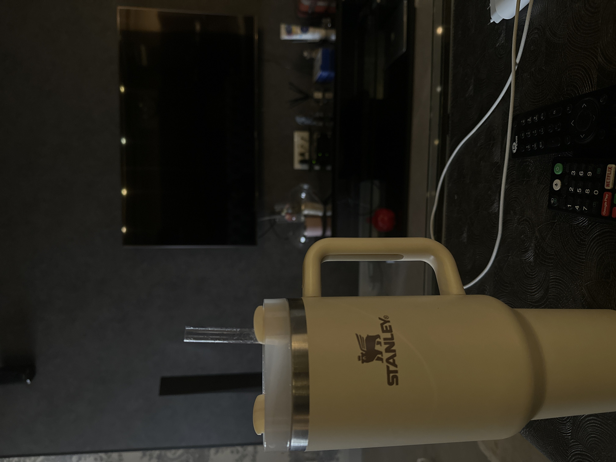 The image shows a cream-colored Stanley brand tumbler with a straw on a dark, textured surface. In the background, there is a dark gray wall with a large television screen, a media console with various objects on shelves, and two remote controls on the surface next to the tumbler. There is no indication this image was taken in Yerevan, Armenia.