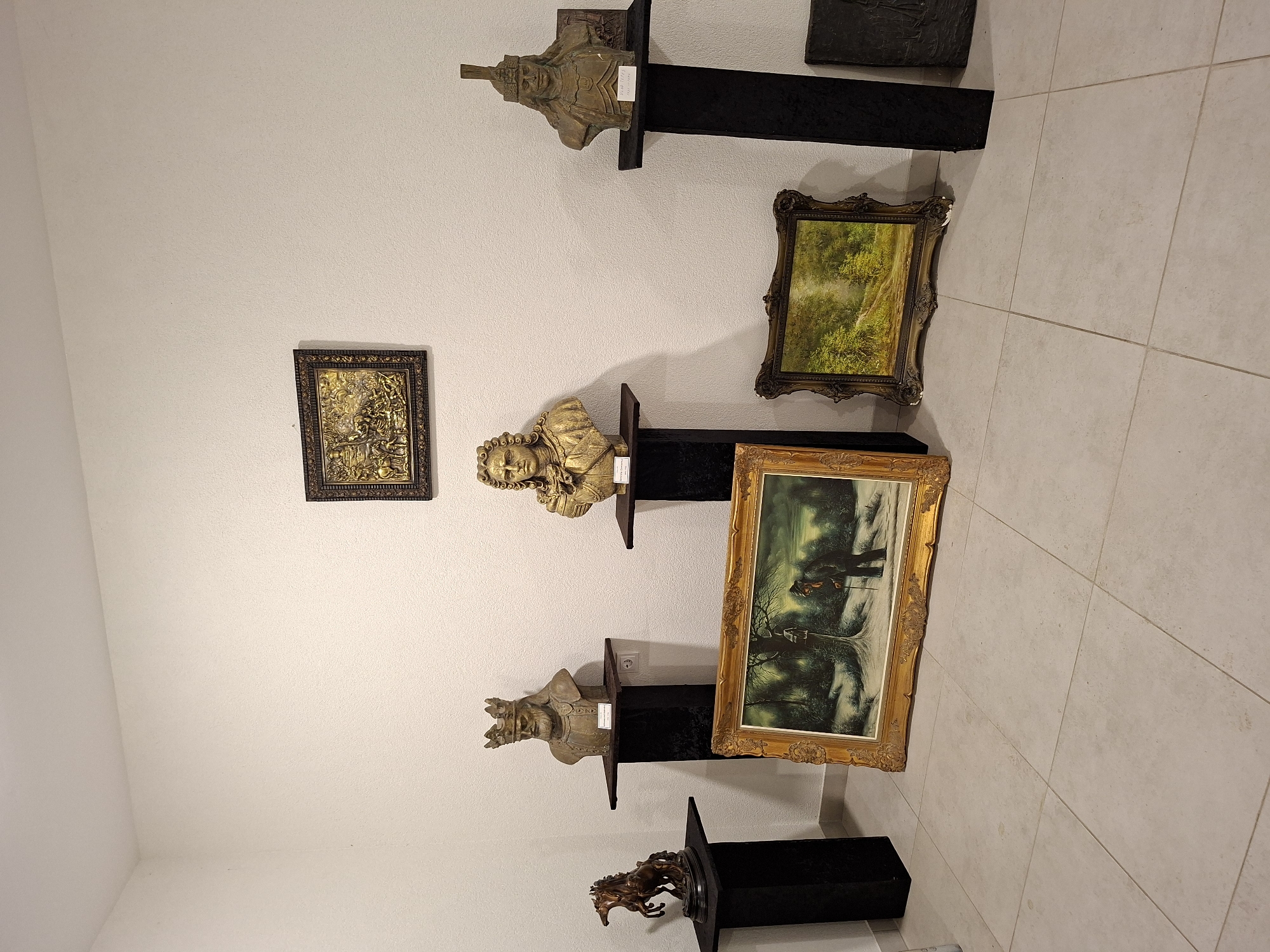 The image depicts an indoor exhibition space, likely a museum or gallery, in the city of Ialoveni, Moldova. The walls are painted white and textured. Several artifacts are displayed on black pedestals and on the floor.
On pedestals from left to right:
* A bronze sculpture of a horse, appearing to be in mid-stride.
* A bust of a man wearing a crown, likely a historical figure, made of a beige material. A label is partially visible below it.
* A golden bust of a man with curly hair and a formal outfit. A label is visible below it.
* A bust of a man wearing a helmet and armor, also made of a beige material. A label is visible below it.
* To the far right, partially visible, is another artifact on a black pedestal, possibly a relief sculpture.
On the floor, from left to right:
* A large framed painting depicting a winter landscape with a figure walking with a cane. The frame is ornate and golden.
* A smaller framed painting of a lush green forest scene. The frame is also ornate and golden.
* A dark rectangular artifact, possibly another relief sculpture or plaque.
The floor is tiled with light-colored square tiles. The overall impression is that of a curated exhibition of historical and artistic pieces.