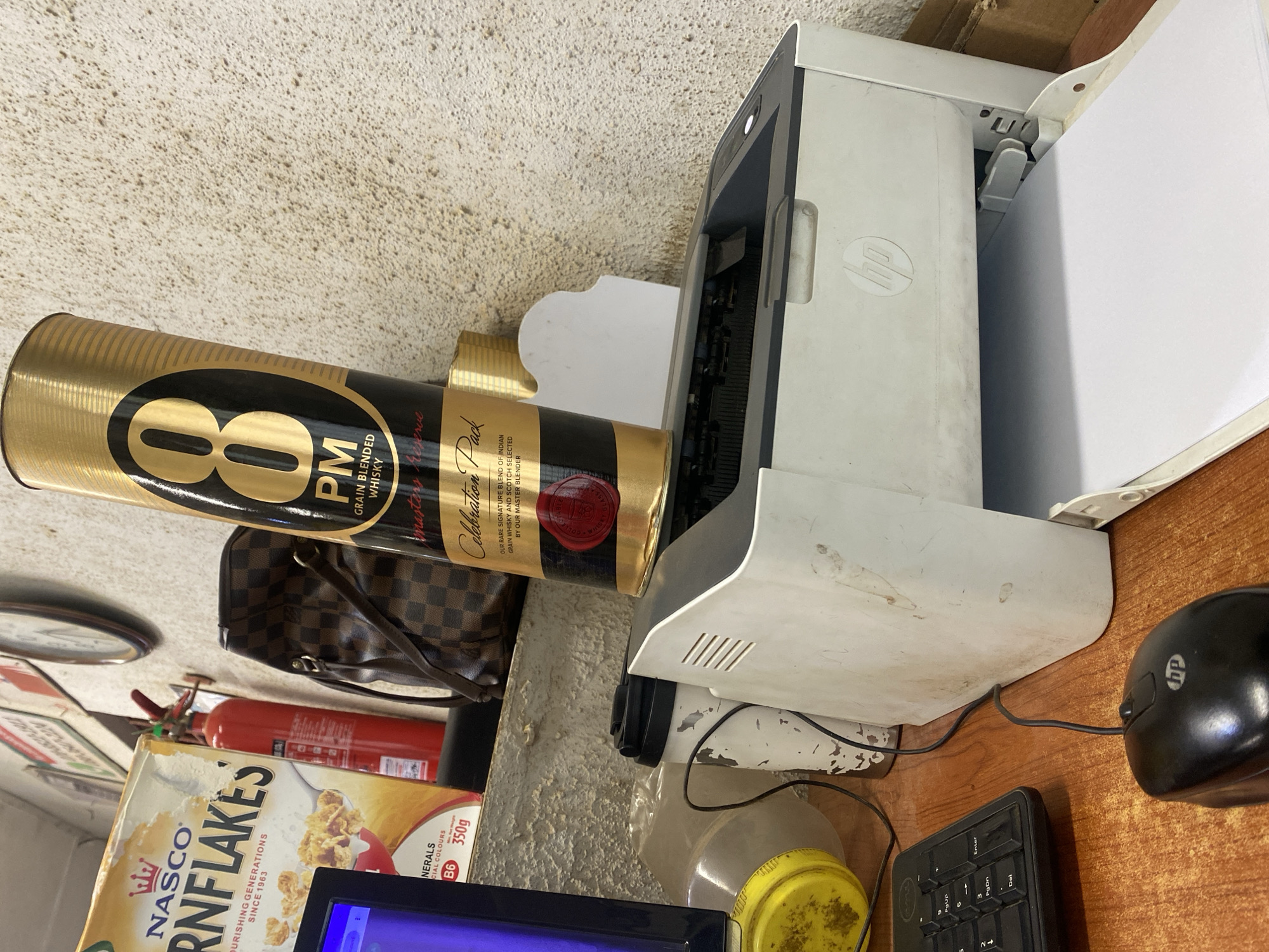 The image depicts a cluttered desk scene, likely in a commercial or residential setting, with an HP printer centrally placed. On the printer, a tall, gold and black cylindrical bottle of "8 PM Grain Blended Whisky" is positioned precariously. To the left of the bottle and printer, a box of "Nasco Cornflakes" is visible, alongside a red fire extinguisher mounted on a shelf. Behind the printer, a brown checkered designer handbag is placed on the same shelf. The desk itself is made of wood and holds a black computer keyboard and a black HP computer mouse. A portion of a computer monitor, displaying a blue screen, is visible on the far left. The background wall is textured and off-white, and a clock is mounted on it, though its face is partially obscured. The scene suggests an environment where various items have been gathered without particular organization, possibly indicating a workspace or storage area. The city of Ikorodu, Nigeria, is the specified location.