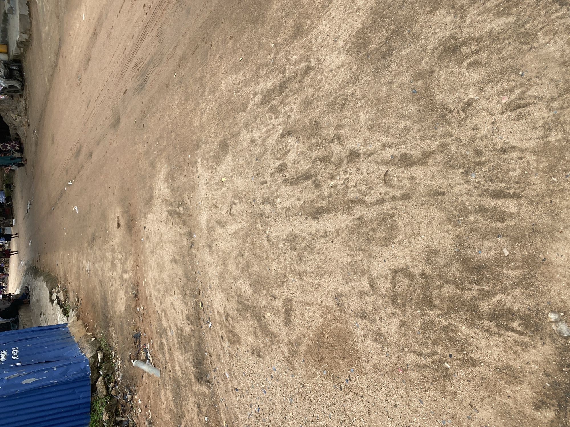The image depicts an outdoor scene, likely a street or pathway in Jalingo, Nigeria. The ground is predominantly sandy and dusty, with visible tire tracks indicating vehicular activity. Scattered debris, including small pieces of plastic and paper, litters the surface.
To the left of the frame, a blue corrugated metal structure, possibly a shipping container or a temporary shelter, is partially visible. Some faint white text is visible on its side, which appears to be "MANKAI" and "M. 1818325". Adjacent to this structure, a rocky embankment with some sparse green vegetation is present.
In the background, along the left edge of the image, a few people are visible, dressed in casual clothing. Their presence suggests a populated area, possibly a market or a residential street. One person is seated near the embankment. The overall impression is of a busy, perhaps unpaved, thoroughfare in a developing area.
The lighting suggests it is daytime, with the sun casting some shadows, but the exact time of day is not precisely determinable. The weather appears to be dry, given the dusty nature of the ground. No specific emotional cues are evident, and the scene conveys a sense of everyday life in this location.
