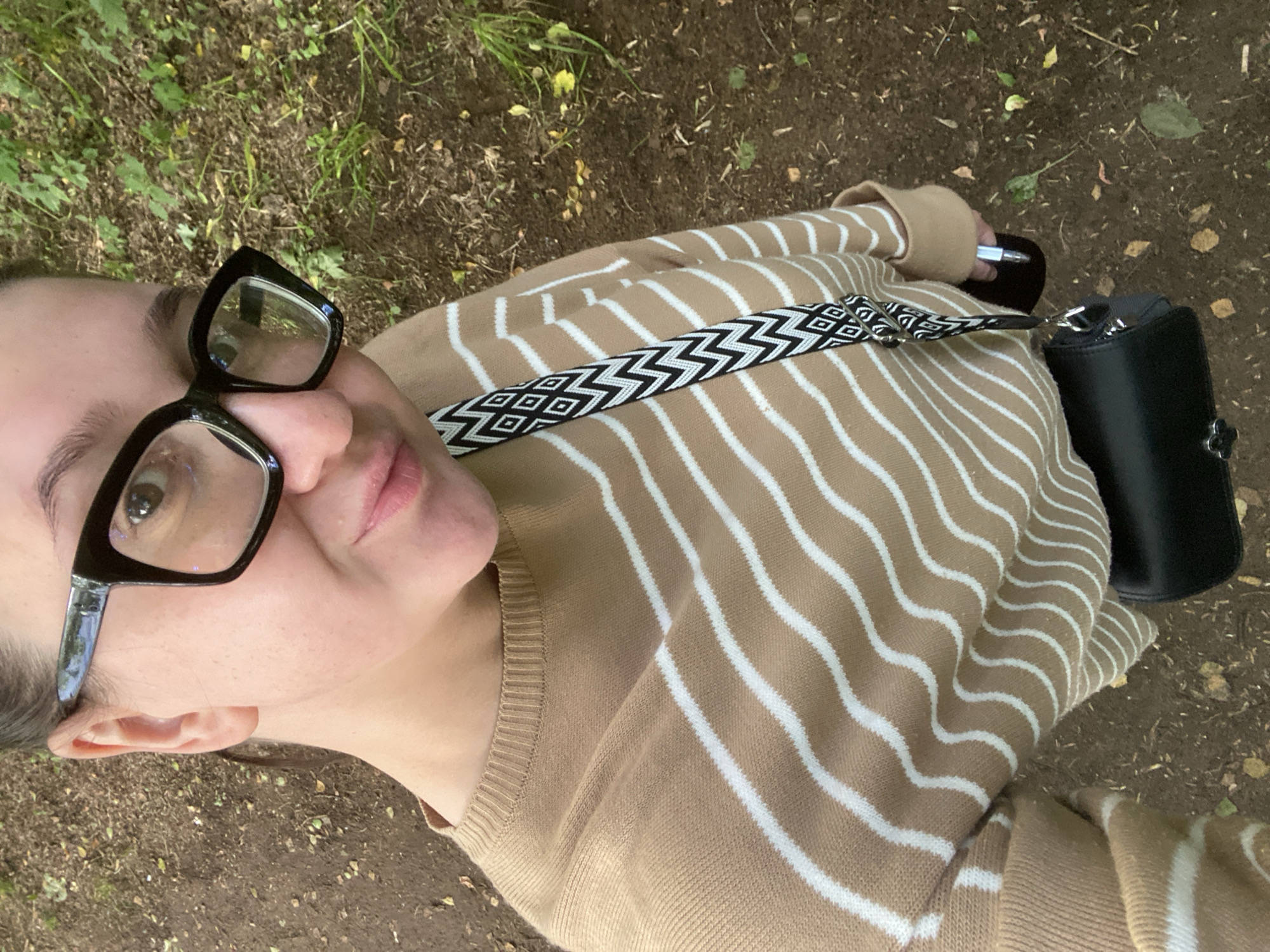 A woman is taking a selfie outdoors. She is wearing a tan and white striped sweater, black glasses, and a black purse with a patterned strap. She is standing on a dirt path surrounded by green foliage. The location is the city of Chaikovsky, Russia.
