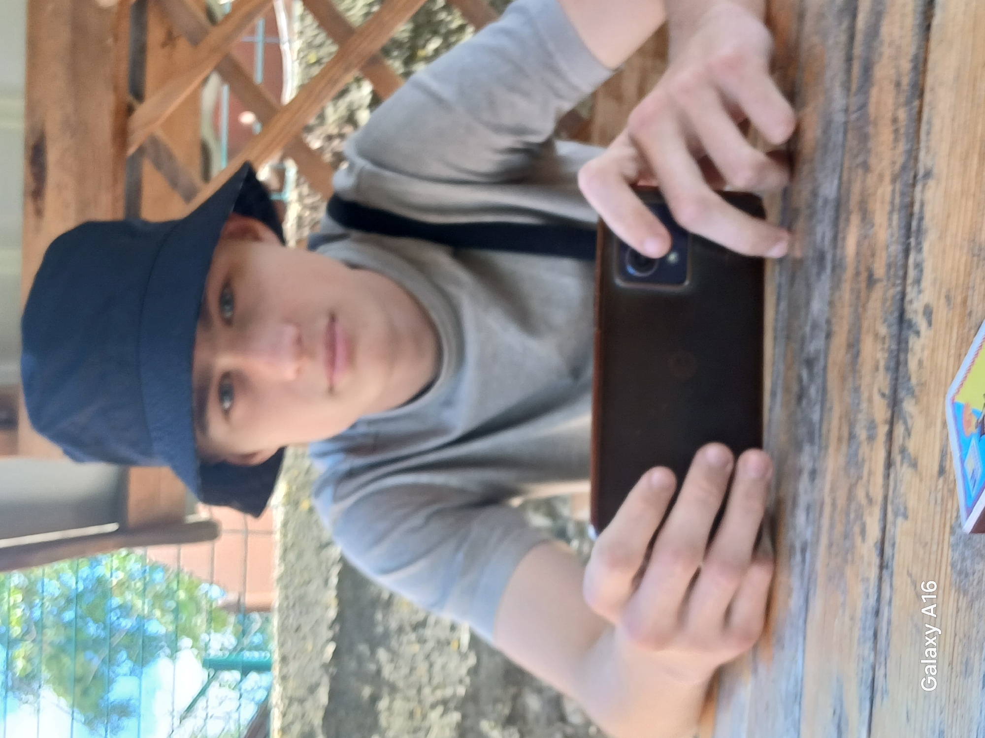 A young male wearing a black bucket hat and a grey t-shirt is sitting at a wooden table outdoors. He is holding a dark-colored smartphone in both hands. The background consists of a lattice structure made of wood, some greenery, and a partially visible brick wall. The lighting suggests it is daytime. The image contains a watermark in the lower left corner that reads "Galaxy A16". The scene appears to be in Ustyvytsia, Ukraine, based on information not present in the image itself.