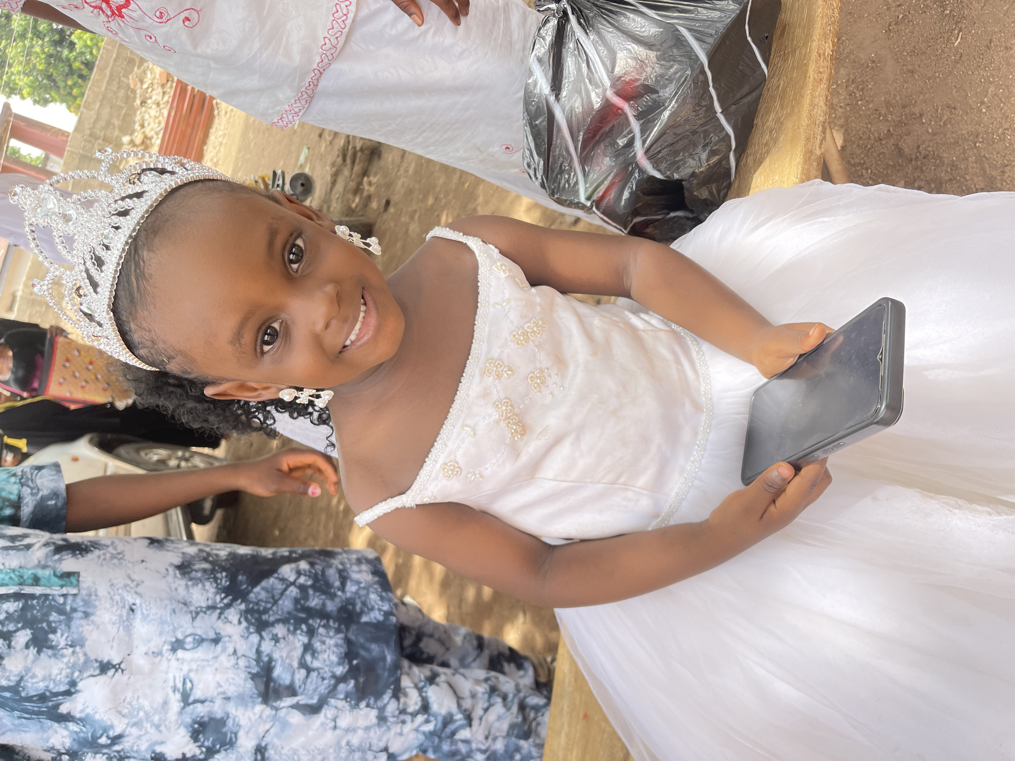 A young girl with dark hair and skin stands outdoors in Akwanga, Nigeria, wearing a white dress, a silver crown, and silver earrings. She is holding a black smartphone. An adult wearing a blue patterned garment is standing nearby on the left. Other partially visible people and a black plastic bag are also present. The ground appears to be dirt.