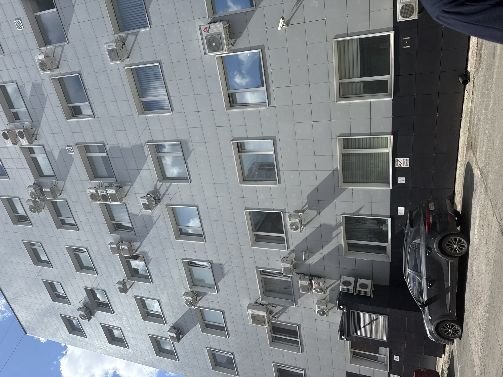 The image shows the side of a modern building with a gray, tiled facade, interspersed with numerous windows and air conditioning units. The building's architecture is utilitarian, with clean lines.
Several air conditioning units, both individual and clustered in pairs or triplets, are mounted on the exterior wall, indicating a functional purpose for the building. The windows vary in size and some reflect the sky and clouds, suggesting it is daytime. The weather appears to be sunny with some clouds.
On the lower right of the image, a dark-colored car is parked next to the building, which has a section clad in black tiles. There is also a portion of what appears to be asphalt or a paved surface next to the car.
Some text is visible on a sign attached to the black-tiled section of the building, but it is too small and pixelated to be deciphered. Another small sign with the number "37" is also visible near the bottom. The presence of a security camera high on the building suggests it is a commercial or institutional space. The overall impression is a contemporary urban environment, likely an office building or apartment complex. The scene is viewed from a slightly elevated perspective, looking upwards at the building.