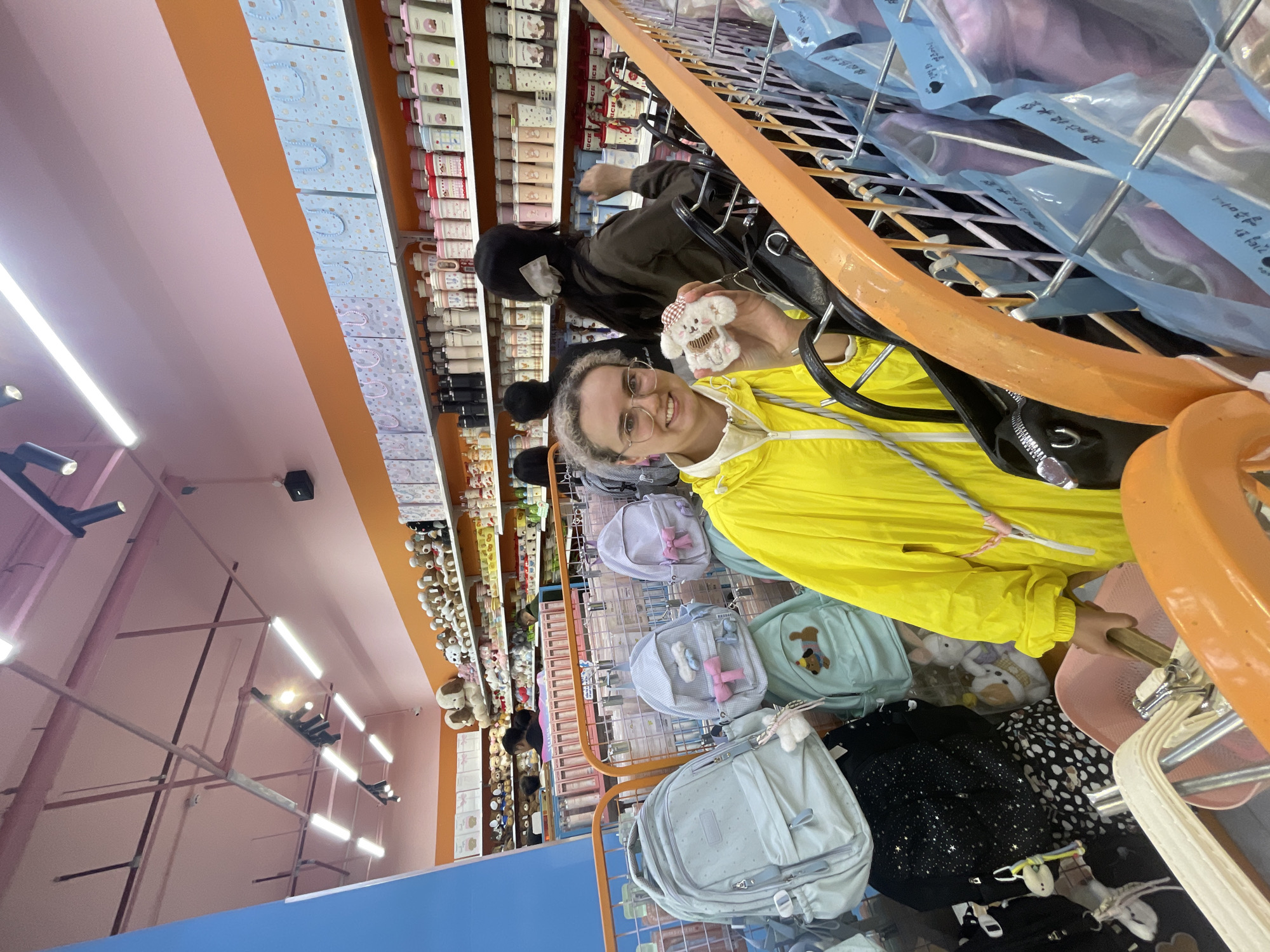 A photo shows a woman wearing a yellow raincoat and glasses holding a small, fluffy toy with a teddy bear head and a cupcake-shaped body. Behind her, there are shelves stocked with various goods, including thermoses and backpacks. Another person, wearing a dark jacket and a face mask, is visible near the shelves. The environment suggests a retail setting or a store. Based on the appearance of the items and the retail setting, it is probable that the photo was taken in Da Lat, Vietnam, due to the popularity of such retail environments in the city.