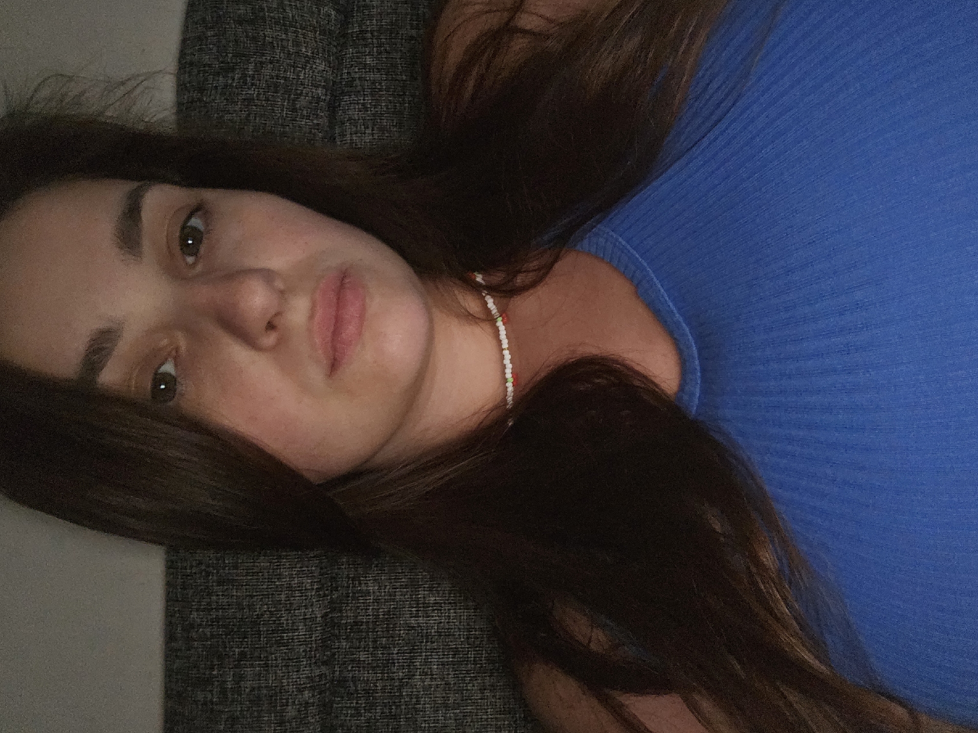 A young woman with long, dark brown hair and light skin is pictured in a close-up shot. She is wearing a blue ribbed tank top and a beaded necklace. Her expression is neutral, and she is looking directly at the camera. The background is a textured gray surface, possibly a couch or upholstery. The lighting is dim, suggesting the photograph was taken indoors in the evening. There is no discernible context for the location within Petropavlivska Borshchahivka, Ukraine, as the image is solely focused on the woman's face and upper body.