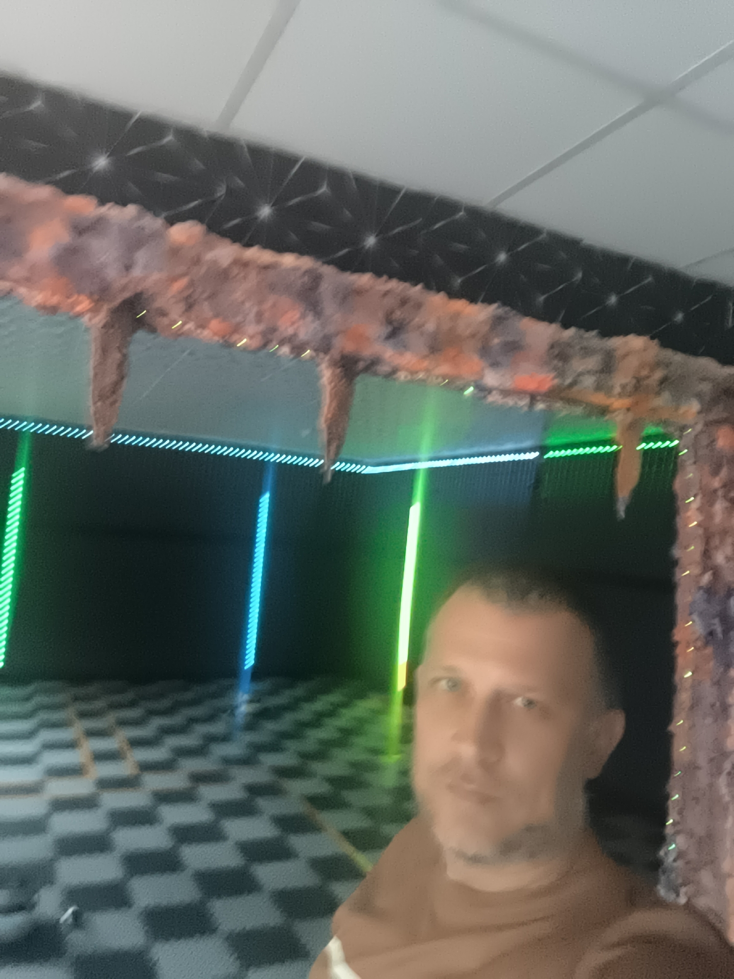 An eye-level indoor shot captures a man partially visible on the right, taking a selfie or being photographed within a uniquely lit and decorated space. The man has short hair and a beard, wearing a light brown or tan shirt, with a neutral expression.
Overhead, a textured, organic-looking beam or archway, adorned with small yellowish lights, stretches across the frame. Several stalactite-like structures hang from this beam. Above it, a dark surface features a reflective, geometric star or snowflake-like pattern, topped by a white drop ceiling with visible grid lines.
The background is dominated by a dark wall, illuminated by vibrant vertical and horizontal neon strip lights in electric green and blue. These lights cast a distinct glow on the environment, creating a futuristic or club-like atmosphere. The floor features a glossy, black and white checkered pattern, reflecting some of the light from the neon strips.
The overall scene suggests an entertainment venue, a themed room, or an art installation, characterized by its distinctive lighting and decorative elements. There is no visible text. The location is Trastenik, Bulgaria.