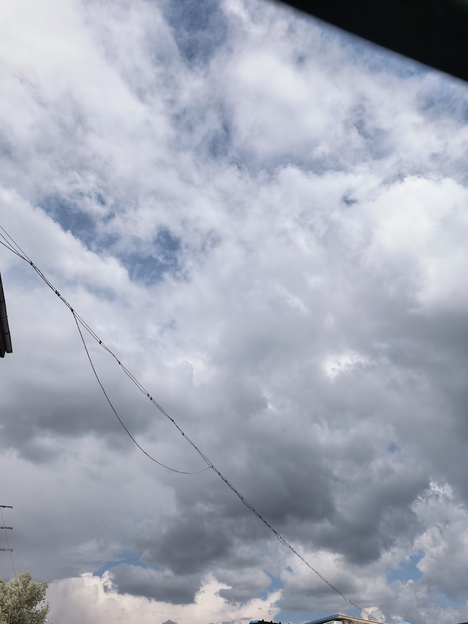 The image depicts a cloudy sky above an urban area. Visible are overhead power lines stretching across the frame, and glimpses of buildings beneath the clouds. The sky is filled with a mix of white and gray clouds, with patches of blue visible in places. The location is described as being in the city of Unknown, Unknown.