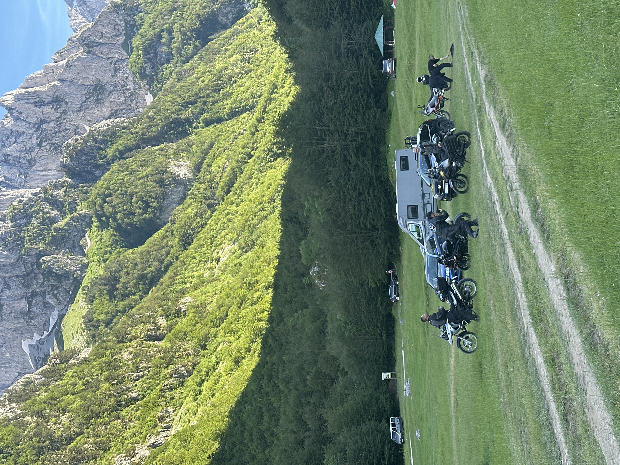 In a grassy meadow at the foothills of the Accursed Mountains in Dolja, Montenegro, a group of motorcyclists have gathered. Several motorcycles, including a white BMW and a black Honda, are parked on the grass. Four individuals, dressed in motorcycle gear, are interacting with the vehicles. One person is standing next to a white motorcycle, another is sitting on a black motorcycle, and two individuals are bent over near another motorcycle. In the background, a grey camper van is parked, and a dark-colored SUV is visible further back. A green-roofed gazebo is situated in the far right of the frame. The surrounding environment is dominated by lush green forest leading up to the steep, rocky slopes of the majestic mountains under a clear blue sky.