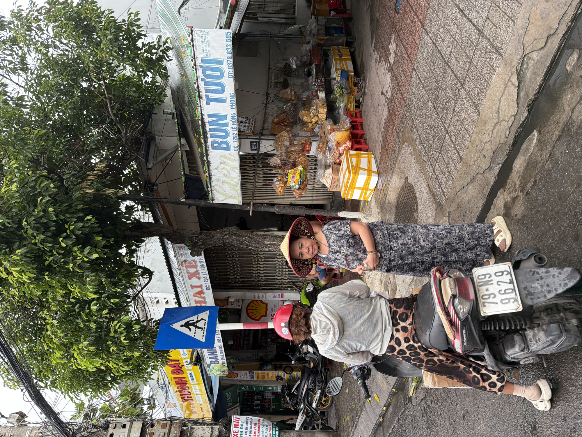 In a street scene in Nha Trang, Vietnam, two women are depicted near a shop. One woman, wearing a red helmet, is seated on a motorcycle, her back to the camera. She has on a light-colored hooded jacket and a distinctive leopard-print skirt. The second woman, facing the camera, wears a conical hat and a dark floral print dress. She is standing, holding what appears to be a small white object in her hands. The motorcycle, a Yamaha, is parked on the street, its license plate reading "79-N9 9629".
Behind the women, a shop displays a variety of goods, including packaged snacks and dry food items. The shop's signage, partially visible, indicates it sells "BUN TUOI" (fresh noodles) and includes a phone number. A blue pedestrian crossing sign is prominently placed on the sidewalk. The street appears wet, suggesting recent rain. The surrounding environment includes trees, buildings with awnings, and other motorcycles parked along the roadside, characteristic of a busy urban area in Vietnam.