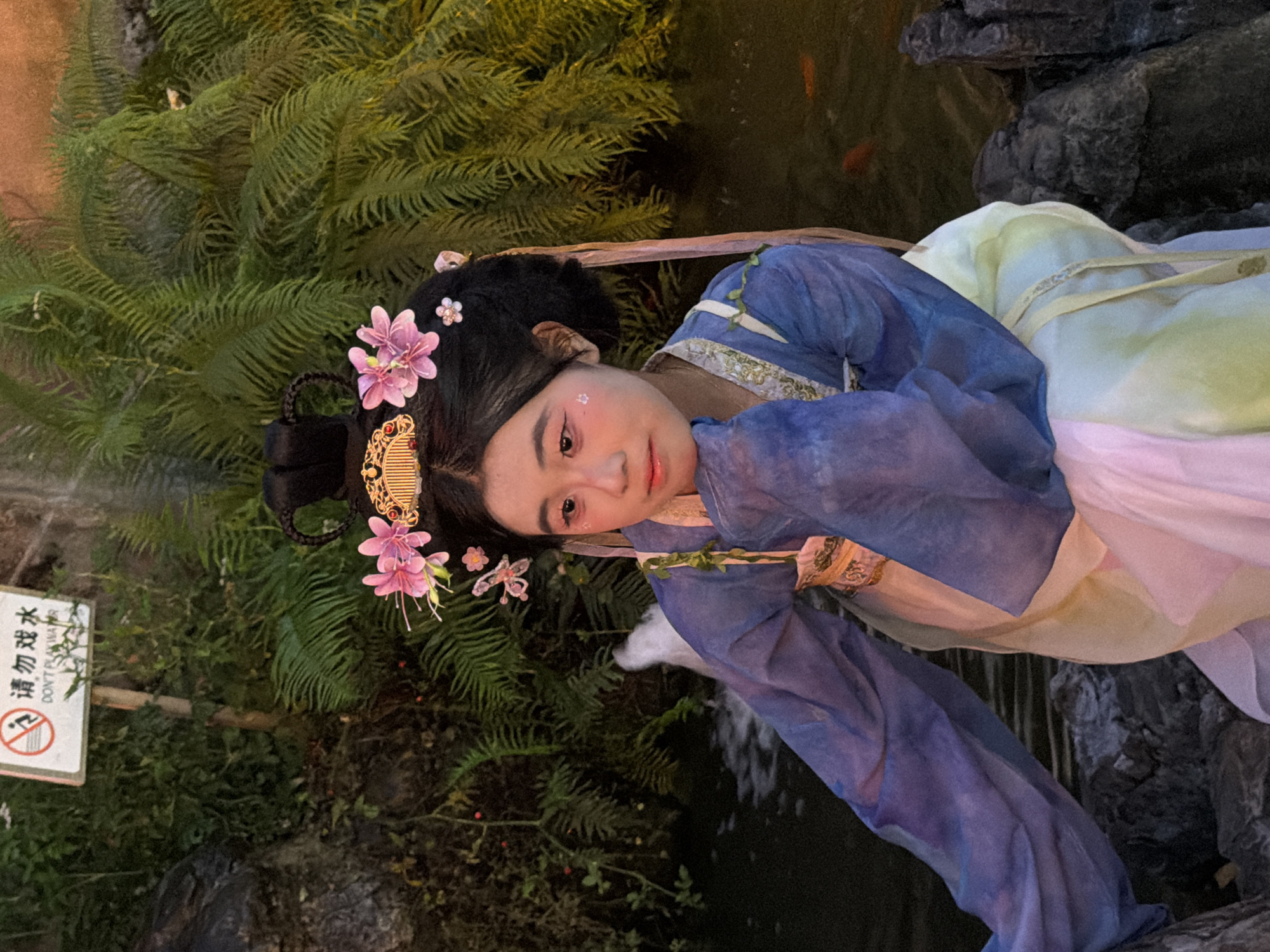 This media file features a young woman dressed in traditional Chinese attire, specifically a flowing blue and purple robe with delicate floral embroidery. Her hair is styled in an intricate updo adorned with pink flowers and decorative hairpins. She is lying down near a body of water, possibly a pond or stream, surrounded by lush green ferns and rocks. The lighting suggests it is evening or nighttime, casting a soft glow on the scene. The woman's gaze is directed towards the viewer, with a serene and perhaps slightly melancholic expression. There are hints of makeup on her face, including subtle blush and what appear to be glittery accents near her eyes. In the background, several small orange fish are visible swimming in the water. A small white sign with Chinese characters and an English translation, "DON'T PLAY WATER," is partially visible on the left side of the frame. The overall scene evokes a sense of tranquility and cultural elegance, set against a natural, watery backdrop. The Yuzhong District in China is provided as a location context.