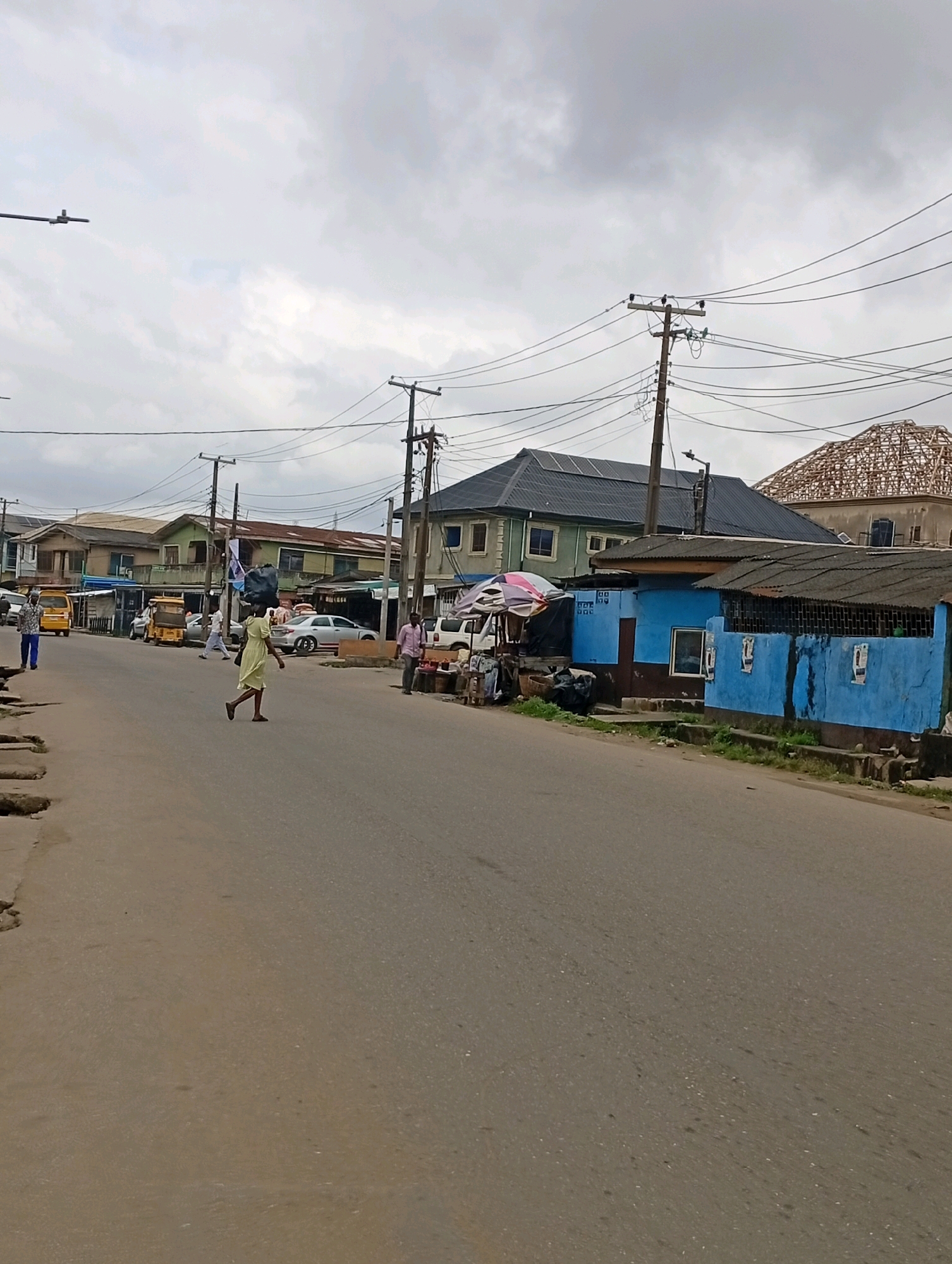 In Akowonjo, Nigeria, on an overcast day, a street scene unfolds. A paved road runs through the middle of the frame, with the left side showing a sidewalk and some concrete debris. Buildings line the right side of the street.
On the road, a woman wearing a yellow dress and carrying a large black plastic bag on her head walks from left to right. To her left, two yellow minibuses are parked, and several other vehicles, including a white car and a silver sedan, are also visible. A man in a white shirt and blue pants walks away from the camera on the far left. Further down the road, a man in a pink shirt walks toward the camera.
The buildings on the right include a structure painted bright blue with a corrugated metal roof. Adjacent to it is a green building with a dark, angled roof, possibly with solar panels. Another building behind it has a partially constructed roof made of wooden beams. Electrical poles with numerous wires are present throughout the scene, strung across the street and between buildings. Various market stalls, some with awnings, are set up along the sidewalk.