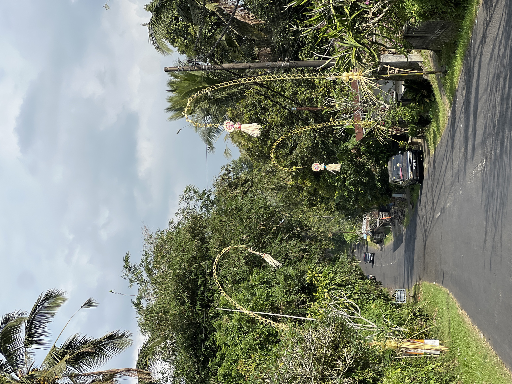 A straight, paved road winds through a verdant, tropical landscape under a cloudy sky. The road is flanked by dense green foliage and tall palm trees. Along the roadside, several decorative, arching structures made of woven straw and bamboo are erected, adorned with tassels and ornate ornaments. A few small, rural buildings are visible nestled amongst the trees. A black SUV is parked on the side of the road, and a dark-colored car is driving away in the distance. The scene appears to be a street in Pupuan, Indonesia, decorated for a local festival or celebration.