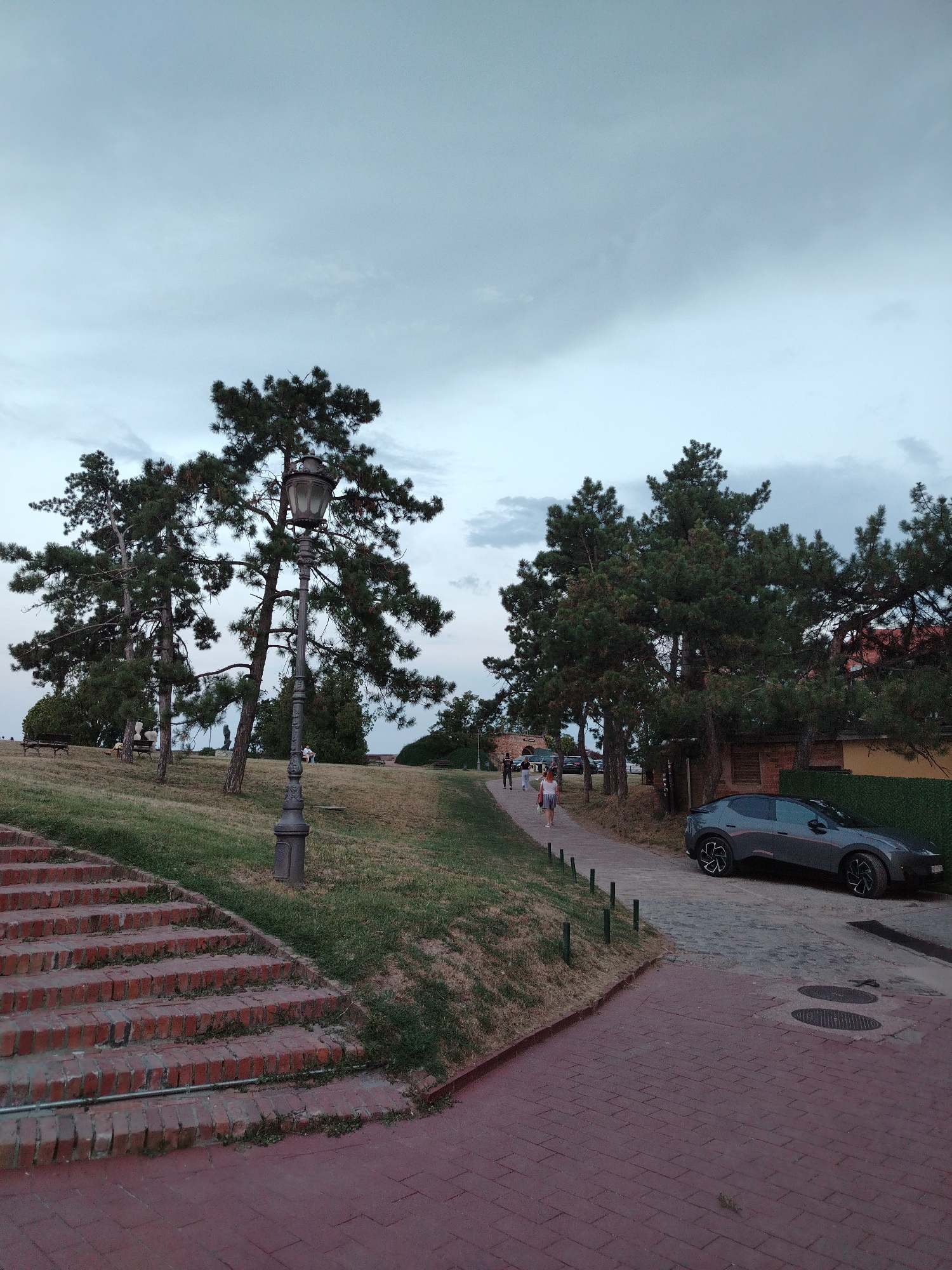 The image depicts an outdoor scene in Petrovaradin, Serbia, likely within the Petrovaradin Fortress. The foreground features a red brick staircase leading up to a grassy slope, with a paved walkway branching off to the right. A vintage lamppost stands prominently on the grassy area. Several pine trees are visible, with some individuals walking along the path in the middle ground. A dark-colored modern car is parked on the paved area near the right side of the image. The sky is overcast and a muted blue. The overall atmosphere suggests a public park or a scenic viewpoint.