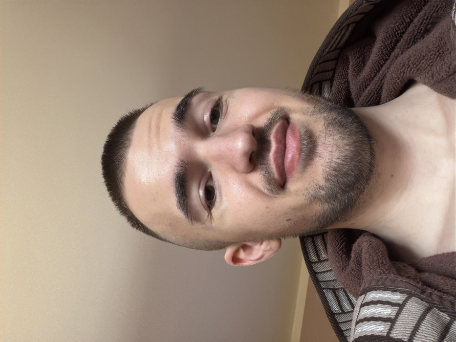 The image displays a close-up, head-and-shoulders portrait of a light-skinned adult male, estimated to be in his 20s or early 30s. He possesses short, dark brown hair cut in a buzz style and a neatly trimmed dark beard and mustache. The subject's eyes are open, and he is looking directly towards the camera with a slight, closed-lip smile. His ears are visible, and there are faint lines around the outer corners of his eyes, consistent with smiling. He is wearing a brown terrycloth robe with a patterned collar featuring horizontal and vertical stripes in light brown and beige tones. The background is a plain, untextured light beige wall, indicating an indoor setting. No specific visual elements within the image content confirm the location as Kladnitsa, Bulgaria; the environment is a generic interior room.