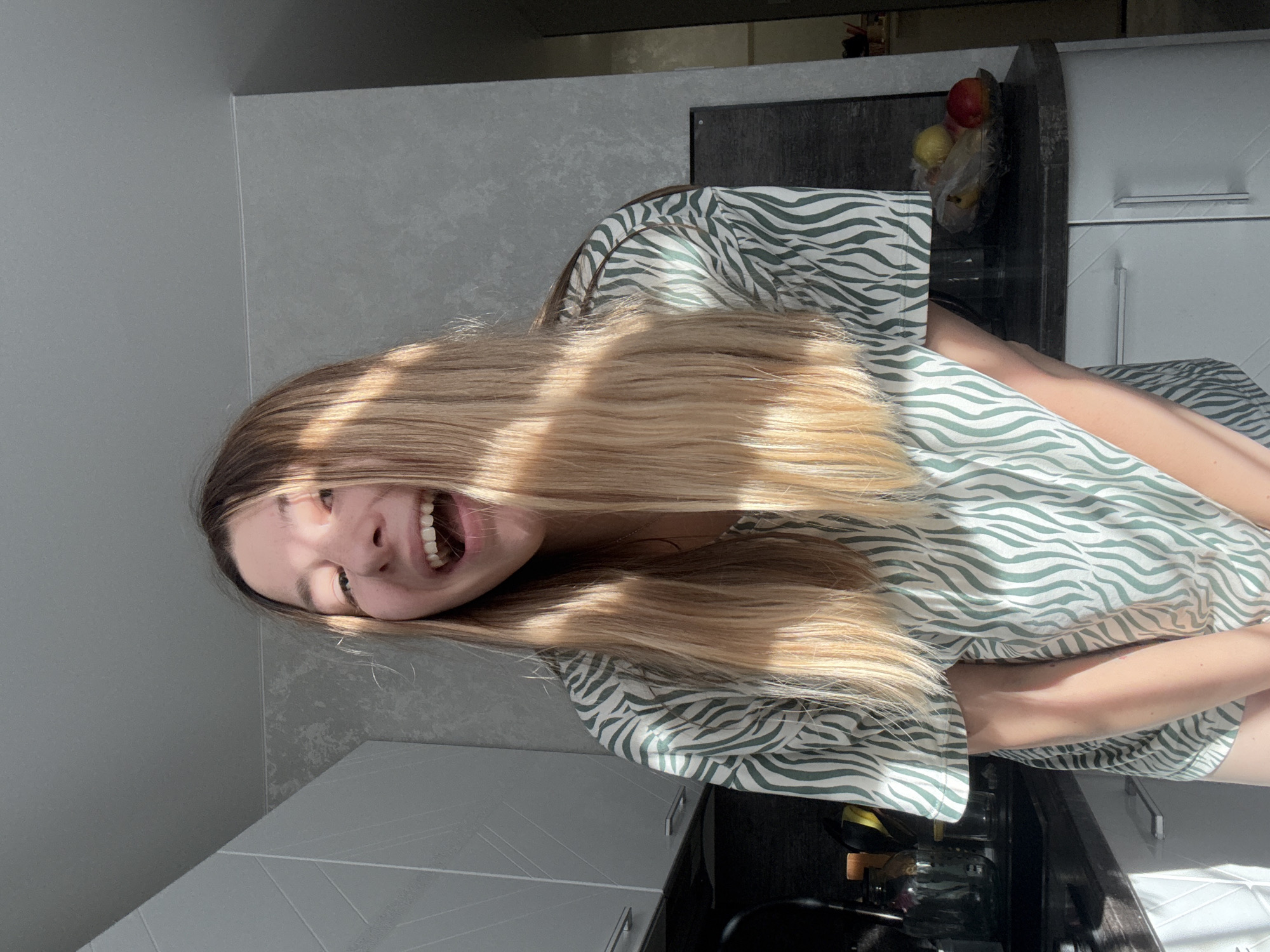 The image shows a young woman with blonde hair and a zebra-print t-shirt, laughing with her mouth open. She is standing in a kitchen, with cabinets and a countertop visible behind her. Sunlight is streaming in from the side, casting shadows across her face and hair. On the countertop, there is a bowl of fruit, including apples and a lemon. The kitchen appears to be modern and well-lit. There is no information to indicate this location is in Moscow, Russia.