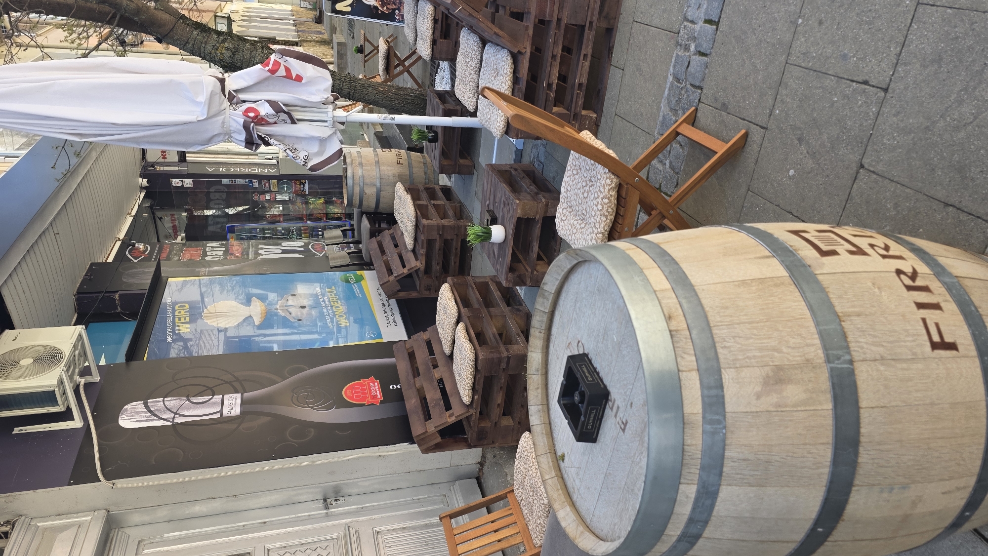 The image shows an outdoor seating area in Blagoevgrad, Bulgaria, with wooden pallets arranged as seating and tables. Wooden barrels are also used as tables, some with ash trays. Cushions are placed on the pallet seating. A store front is visible behind the seating area, with advertisements and an air conditioning unit. The street is paved with grey stones. An umbrella provides shade.