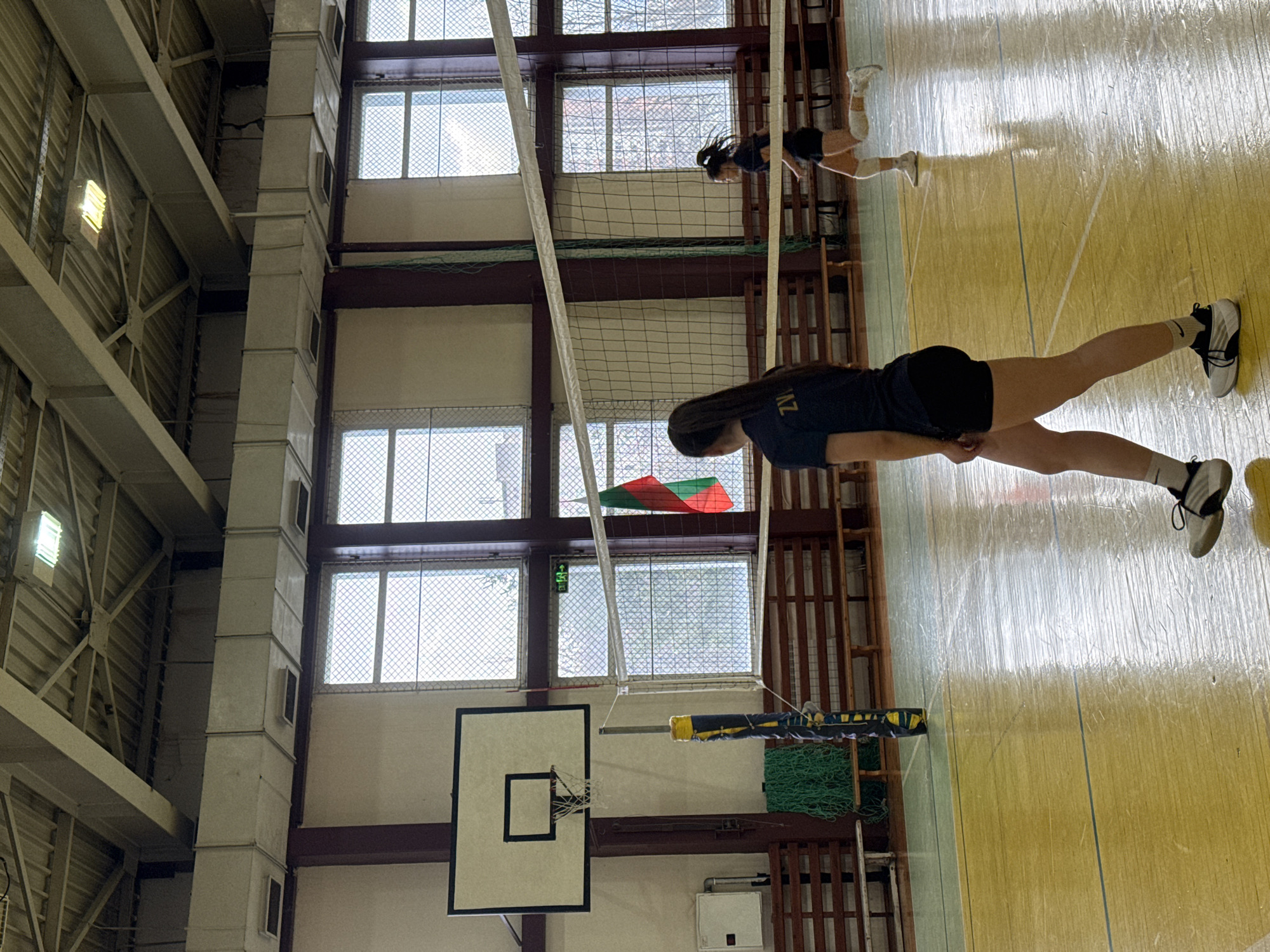 The image shows an indoor volleyball court, likely in a school gymnasium. Two female volleyball players are present. One player is standing near the foreground, wearing a dark blue shirt and black shorts, with white socks and black and white sneakers. The other player is farther away, near the net, also in a dark shirt and shorts. The volleyball net is visible. A basketball hoop is on the back wall. The gymnasium has tall ceilings with industrial-style support beams and several windows. The floor is a glossy light-yellow wood. A Bulgarian flag is visible above the basketball hoop. The location, Sofia, Bulgaria, cannot be ascertained from the image.
