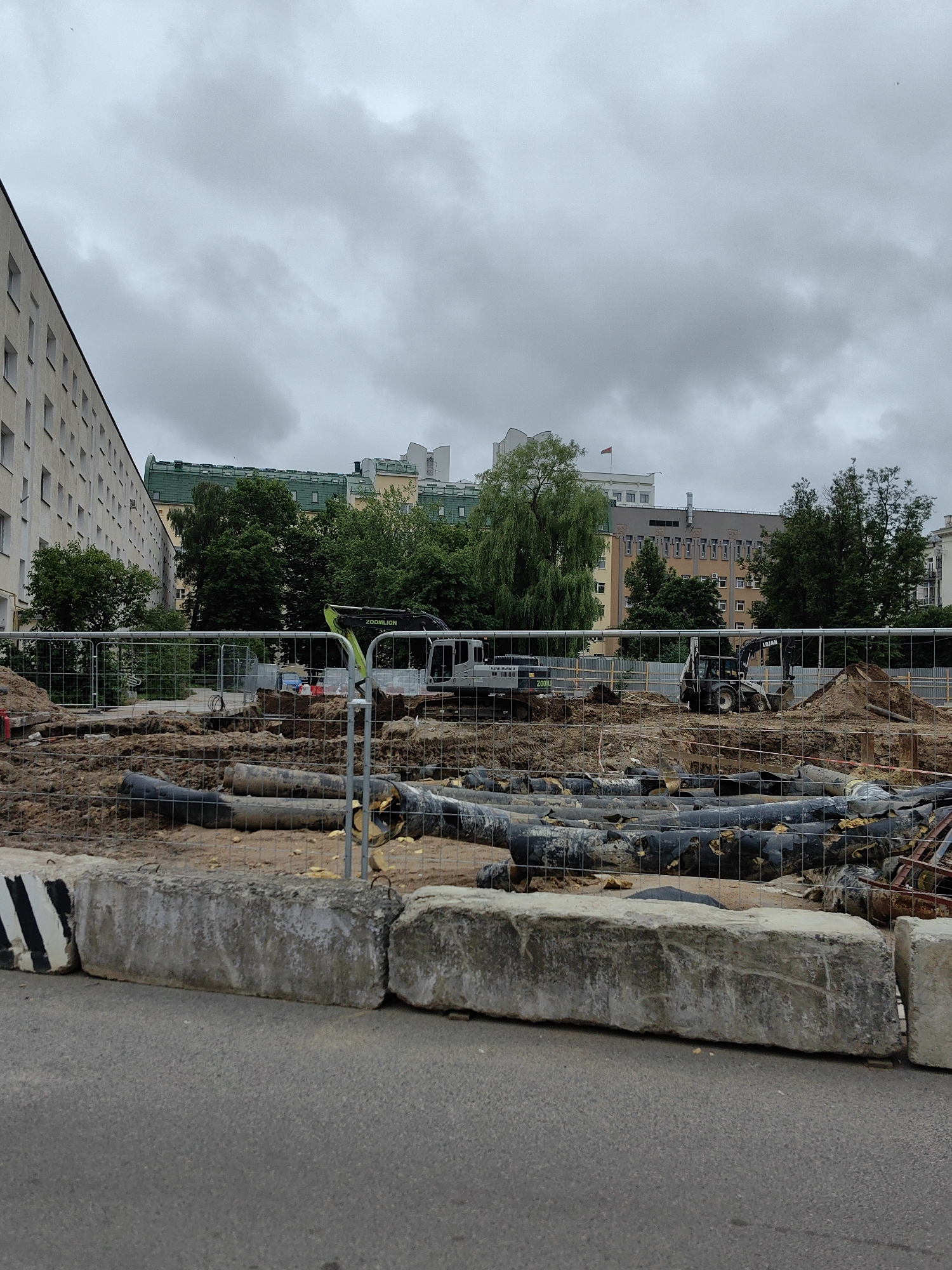 The image shows a construction site in the city of Minsk, Belarus, on a cloudy day. The foreground is dominated by a paved surface and large concrete barriers. Behind these barriers is a fenced-off construction area with exposed earth, piles of black pipes, and construction equipment, including a gray excavator with "ZOONILION" branding and a smaller black loader with "LOIAN" branding. In the background, several multi-story buildings are visible, some with green roofs, and numerous trees. A red flag is visible on one of the buildings. The overall scene depicts an active urban construction project under a gray, overcast sky.
