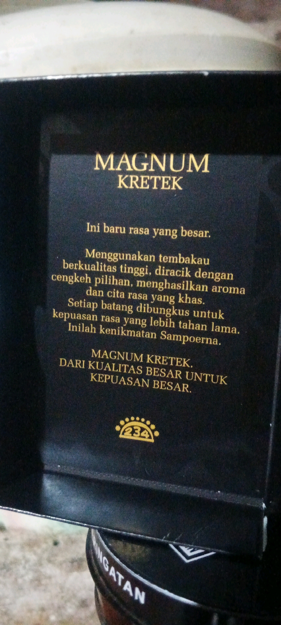 The image shows a close-up of a black box with gold lettering. The text on the box reads "MAGNUM KRETEK" and then a paragraph in Indonesian describing the product. Below this is another line of text that reads "MAGNUM KRETEK. DARI KUALITAS BESAR UNTUK KEPUASAN BESAR." At the bottom of the box, there is a circular emblem with the number "234" inside. The box appears to be made of cardboard. The lighting suggests an indoor or shaded outdoor environment. The lower portion of the image shows the curved edge of what appears to be another object, possibly a container or a base, with white text that is partially visible and reads "INGATAN". The background is out of focus and appears to be a neutral, possibly concrete or stone surface. There are no people visible in the image. The image does not provide any specific information about the city of Penanggungan, Indonesia, nor does it depict any scene within that city. The focus is solely on the product packaging.