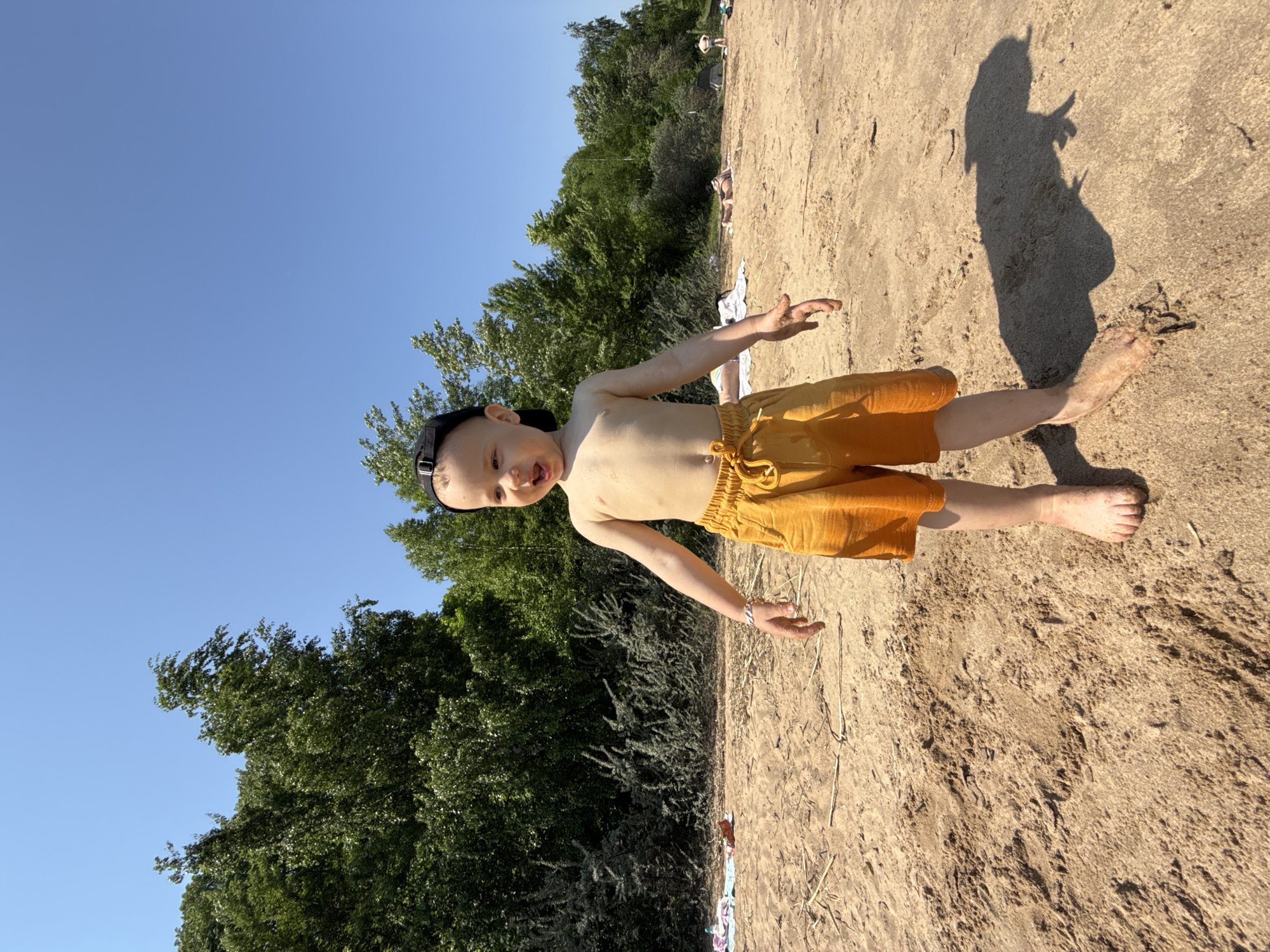 A young child, presumably a toddler, stands on a sandy surface near vegetation. The child is wearing a dark-colored cap and orange shorts. Their arms are outstretched. The background features trees and a clear blue sky. The location may be Rusanovo, Russia, but this cannot be definitively confirmed from the image. There is a shadow cast on the sand adjacent to the child. Other people and objects appear in the background, including what may be towels and other beach items.