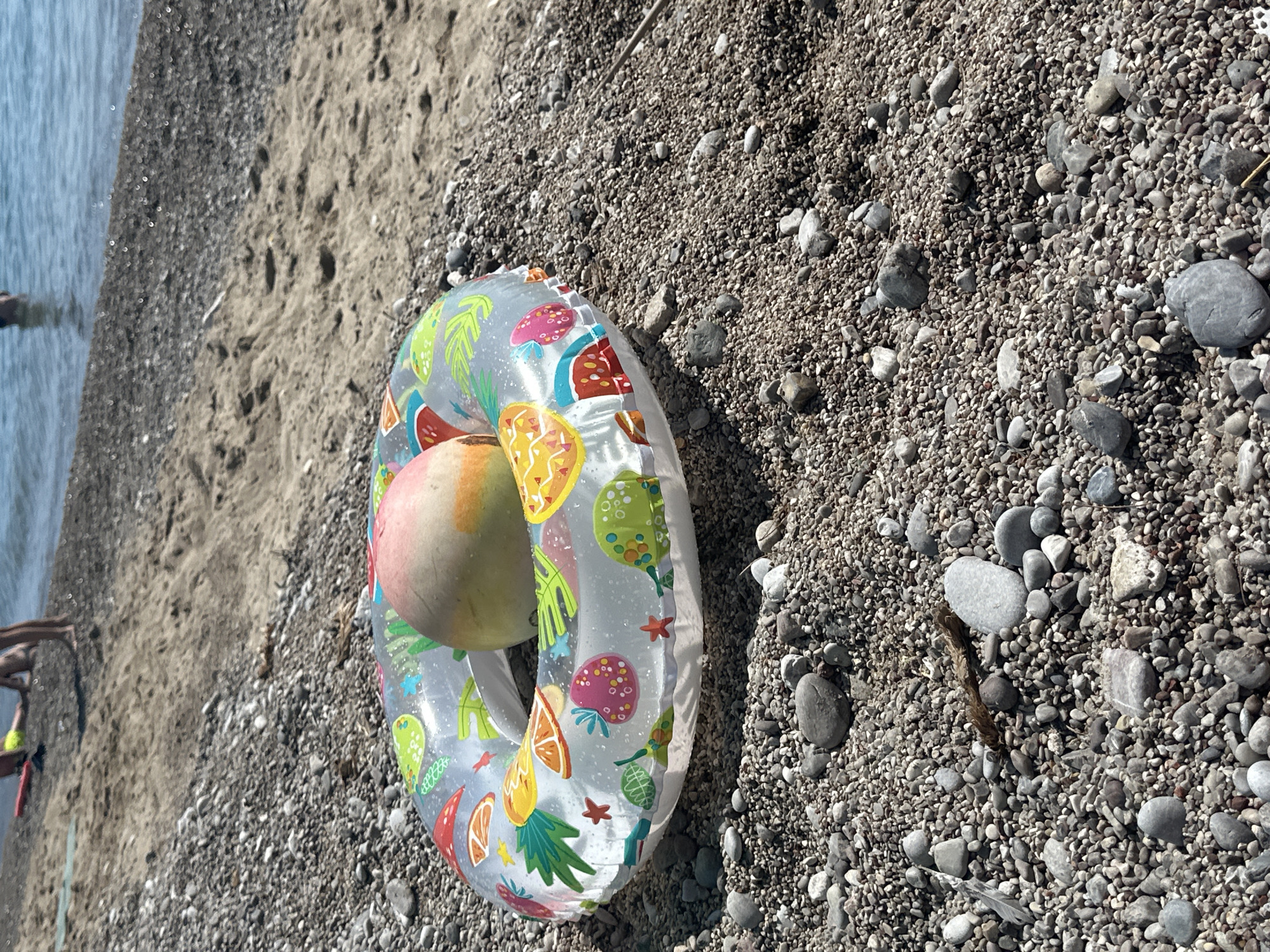An inflatable swim ring lies on a pebble beach near the water. The swim ring is transparent with colorful fruit patterns, including strawberries, watermelons, and citrus slices. A light-colored fruit, possibly a peach or apricot, rests inside the swim ring. The beach is covered in small, grey and brown pebbles. In the background, the blue water of the sea is visible, with the faint figures of people wading in the shallows. The location is Đurovići, Montenegro.