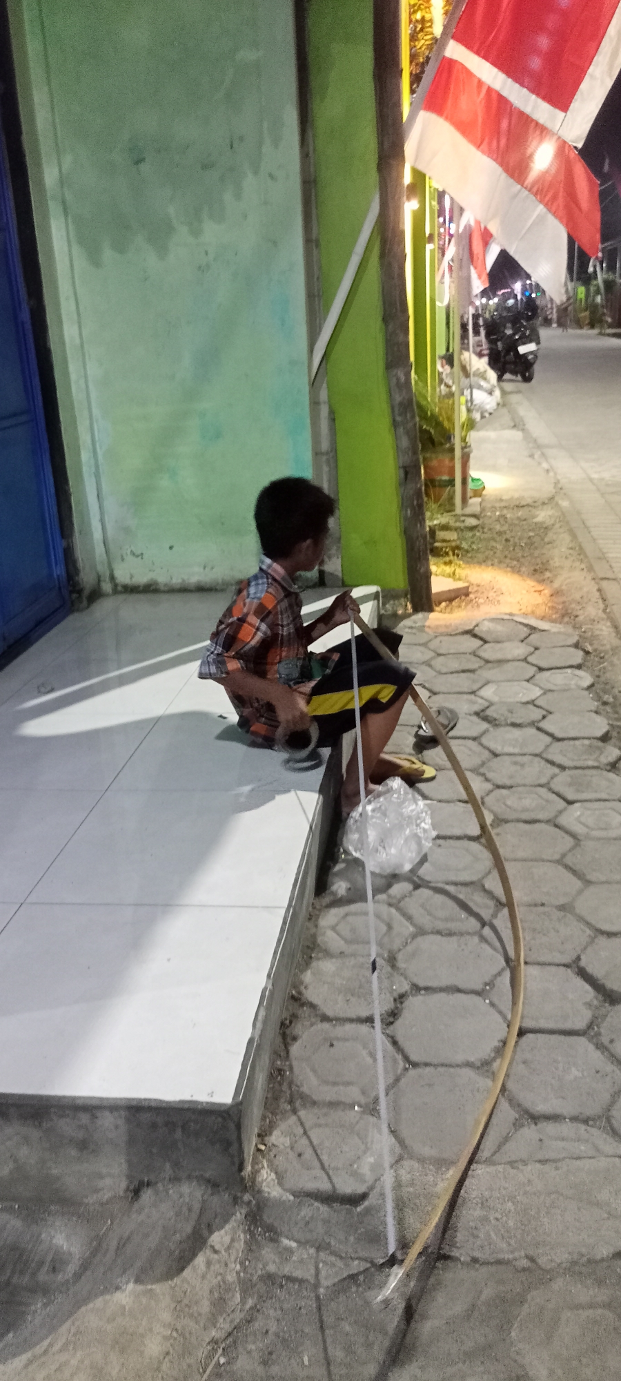 A young boy in a checkered shirt and dark shorts with a yellow stripe sits on a white tiled ledge, facing away from the camera. He is holding a long, thin stick or pole in his hands. To his right, on the ground, is a plastic bag filled with what appears to be crumpled paper or plastic. The boy is positioned on a raised platform next to a paved walkway. The walkway is made of hexagonal concrete blocks and is lined with small plants and some lighting fixtures. A motorcycle is parked on the street in the background. In the upper right of the frame, red and white flags are displayed. The scene appears to be outdoors, possibly on a street in Gempolklutuk, Indonesia, at dusk or night, as indicated by the artificial lighting. The background shows a building with green and light blue walls.