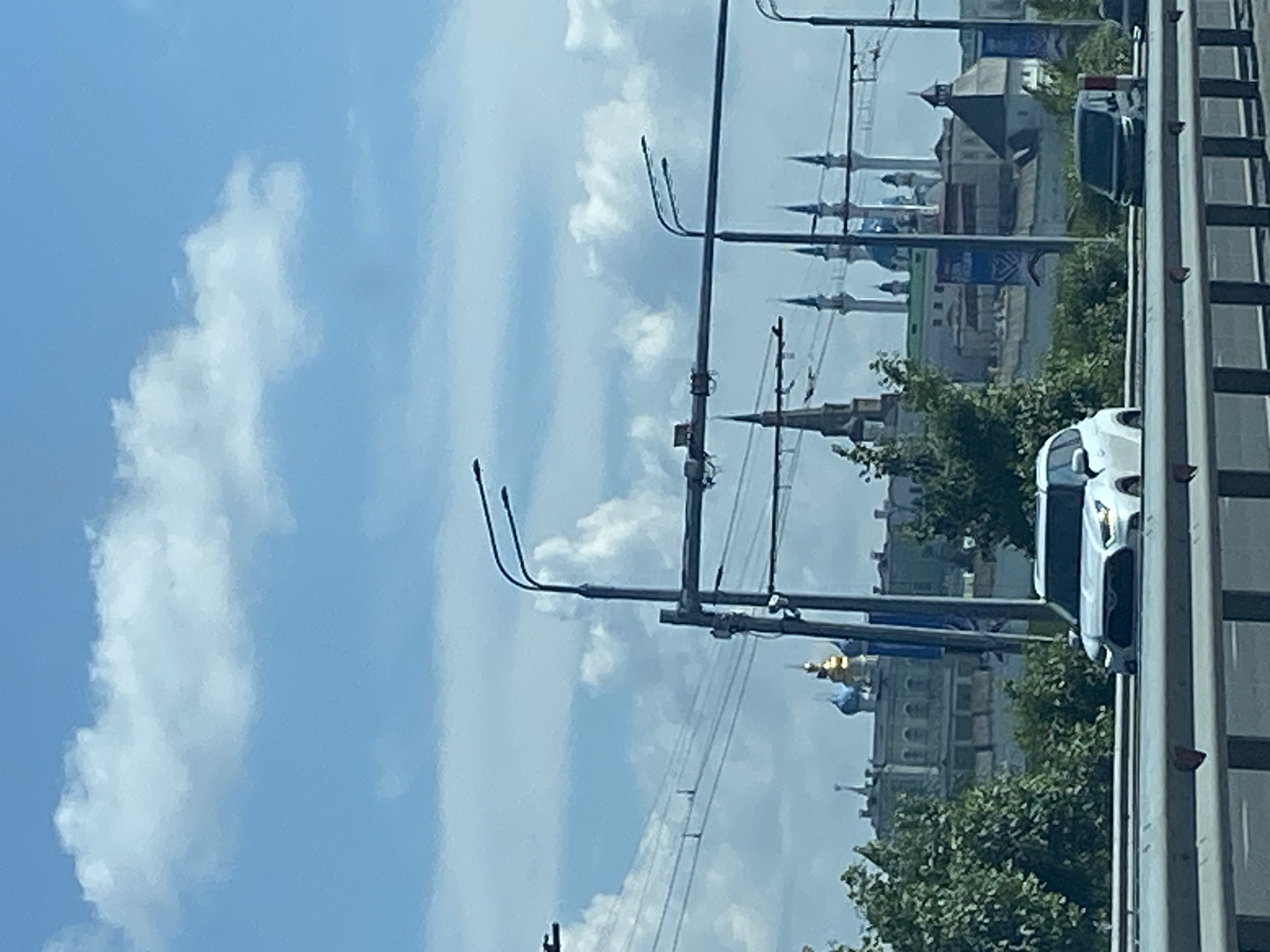The image shows a view of the Kazan Kremlin in Kazan, Russia, from a highway overpass. In the foreground, a white SUV is driving on the highway. Behind the SUV, the Kazan Kremlin complex is visible, with its iconic minarets and domes. Trees and buildings of the city can be seen between the highway and the Kremlin. The sky is blue with some clouds. The overall scene depicts a common view of the historic site from a modern infrastructure.
