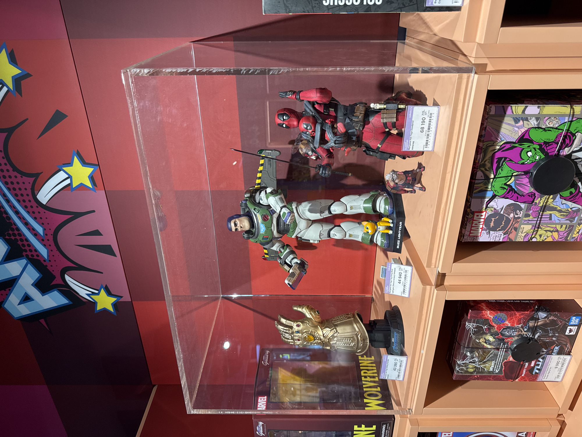 This media file shows a display of collectible action figures in a store, likely a toy shop or a department store in Yekaterinburg, Russia. The figures are prominently displayed in a clear acrylic case.
The primary subjects are action figures of well-known characters from Marvel Comics and Disney Pixar's Toy Story franchise. In the upper part of the display, there are at least two figures of Deadpool, one appearing to be in a dynamic pose. Below Deadpool, there is a detailed action figure of Buzz Lightyear from Toy Story, with a small yellow cat-like companion figure attached to his leg. At the bottom of the display case, a golden gauntlet, resembling the Infinity Gauntlet from Marvel's Avengers, is mounted on a stand, with the word "WOLVERINE" visible on the box beneath it.
Additional boxes of action figures are visible on shelves to the right of the main display. These include figures related to Marvel characters, such as the Green Goblin and Thor.
The background consists of a red wall with comic book-style graphics, featuring stars and stylized "POW!" or "BAM!" elements. The shelves are made of a light-colored material. Price tags are visible on some of the figure boxes, with prices like "68 190", "44 940", "43 390", and "9 990". Text on the price tags and boxes includes Russian and English, with brand names like "Gallery", "Marvel", "Hot Toys", "Bandai", and character names.
The scene appears to be well-lit, suggesting an indoor retail environment. No specific time of day or weather conditions are indicated. The overall impression is one of a retail display aimed at collectors and fans of these popular franchises.