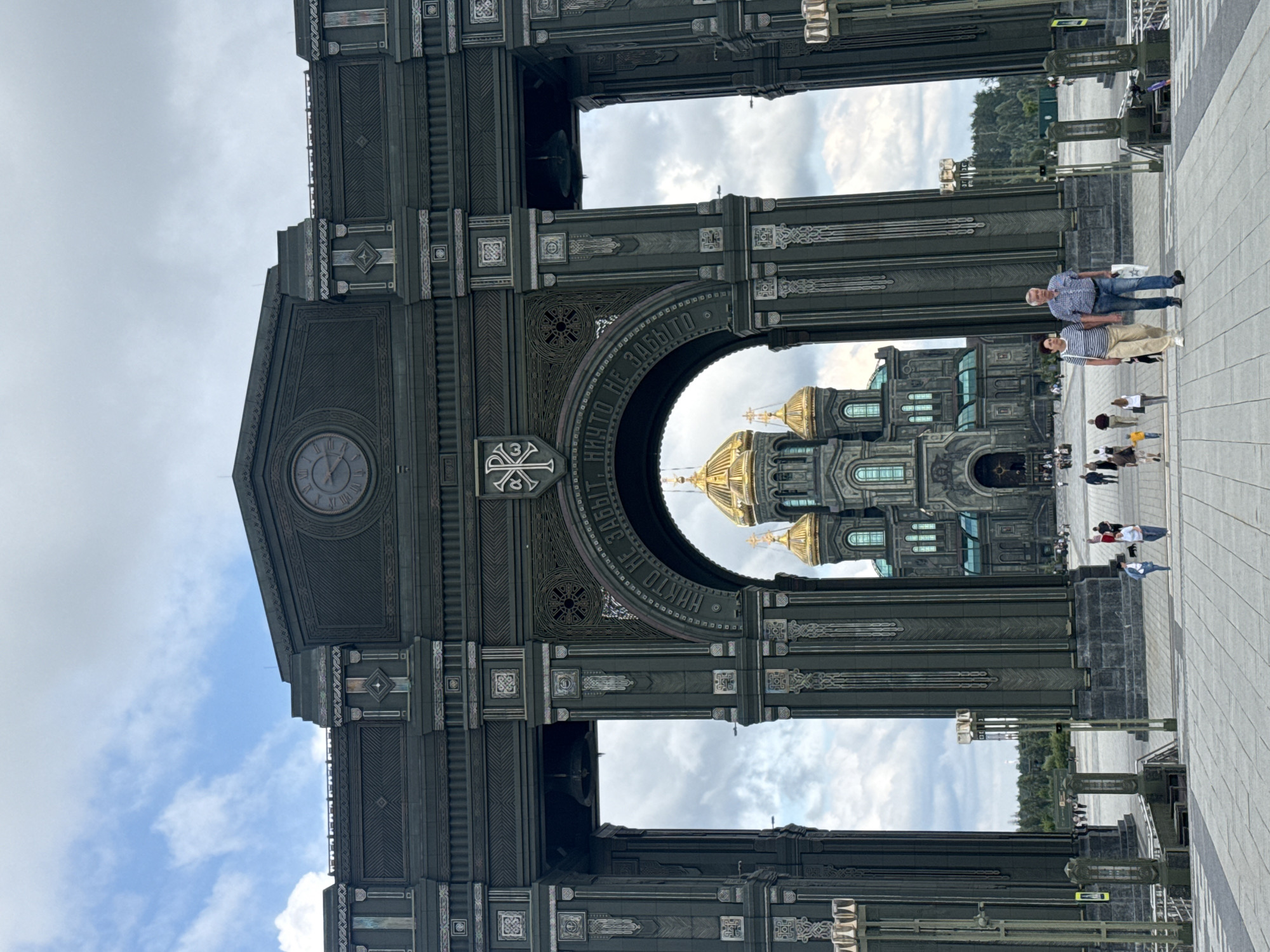 The image depicts the Main Cathedral of the Russian Armed Forces in Zelenaya Roshcha, Russia, framed by a large, ornate archway. The cathedral, with its prominent golden domes, is the central focus, positioned symmetrically through the arch. The archway itself is dark green and features intricate detailing, including inscriptions and decorative patterns. A large clock face is visible at the top of the arch.
In the foreground, a paved pathway leads towards the archway and cathedral. Several individuals are walking on this pathway. Most prominently, a couple, a man and a woman, are walking hand-in-hand towards the viewer. The woman is wearing a striped shirt and light-colored pants, while the man is in a plaid shirt and jeans. Other people, including children, are dispersed in the middle ground and background, also walking on the plaza. The sky above is partly cloudy. The overall scene suggests a public space on a clear day, with visitors exploring the architectural complex.
